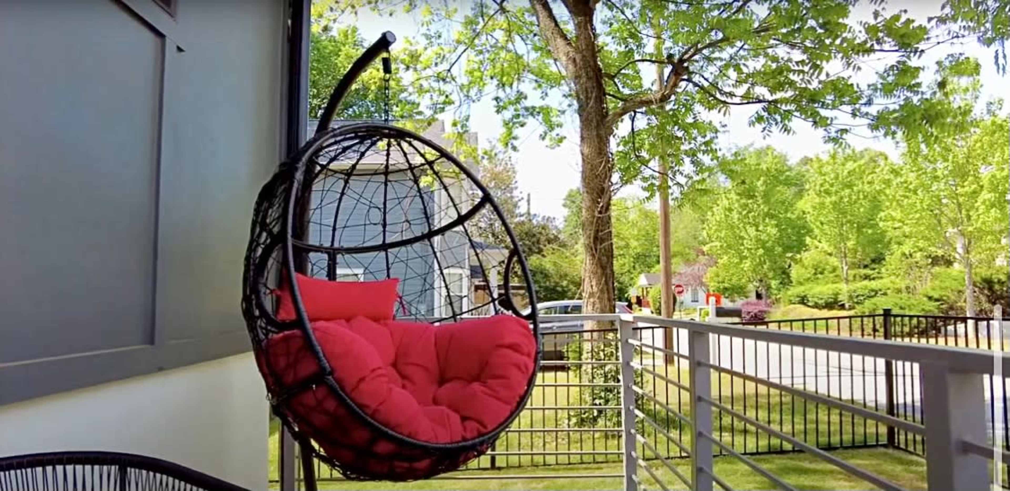 A hanging chair with red cushions is suspended from a balcony surrounded by greenery.