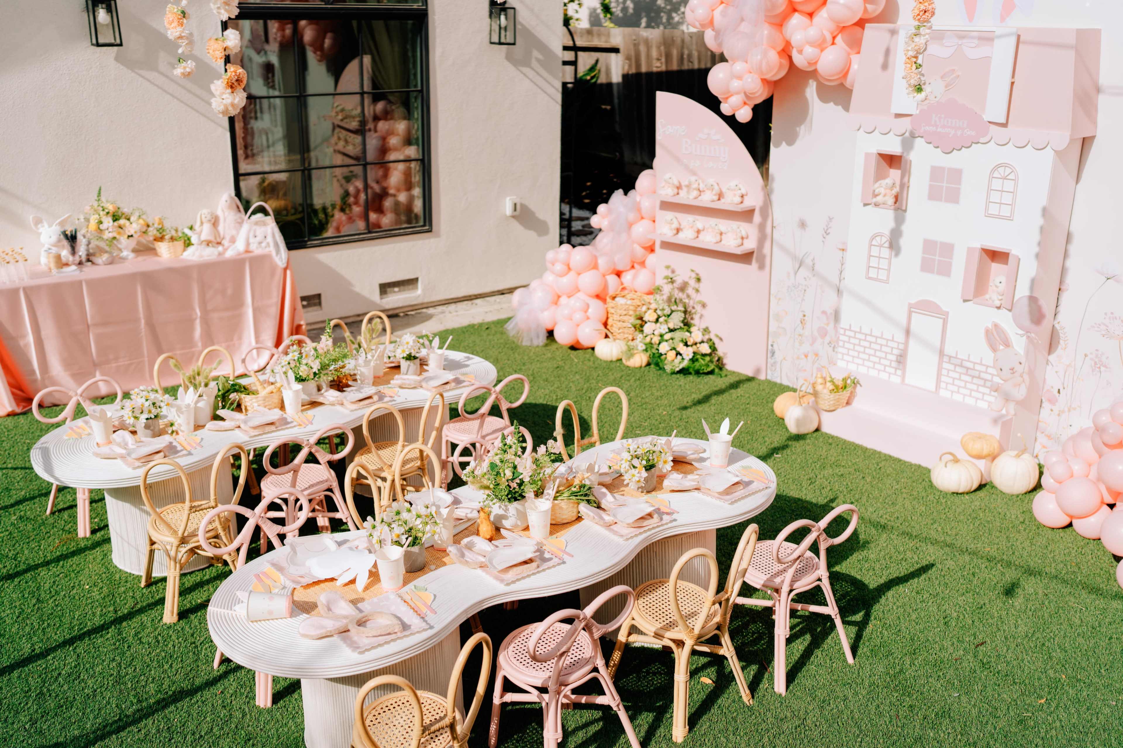 The scene features an outdoor party setup with multiple white tables arranged in a wavy pattern, adorned with pink decor and floral centerpieces, and a backdrop displaying a decorative house design.