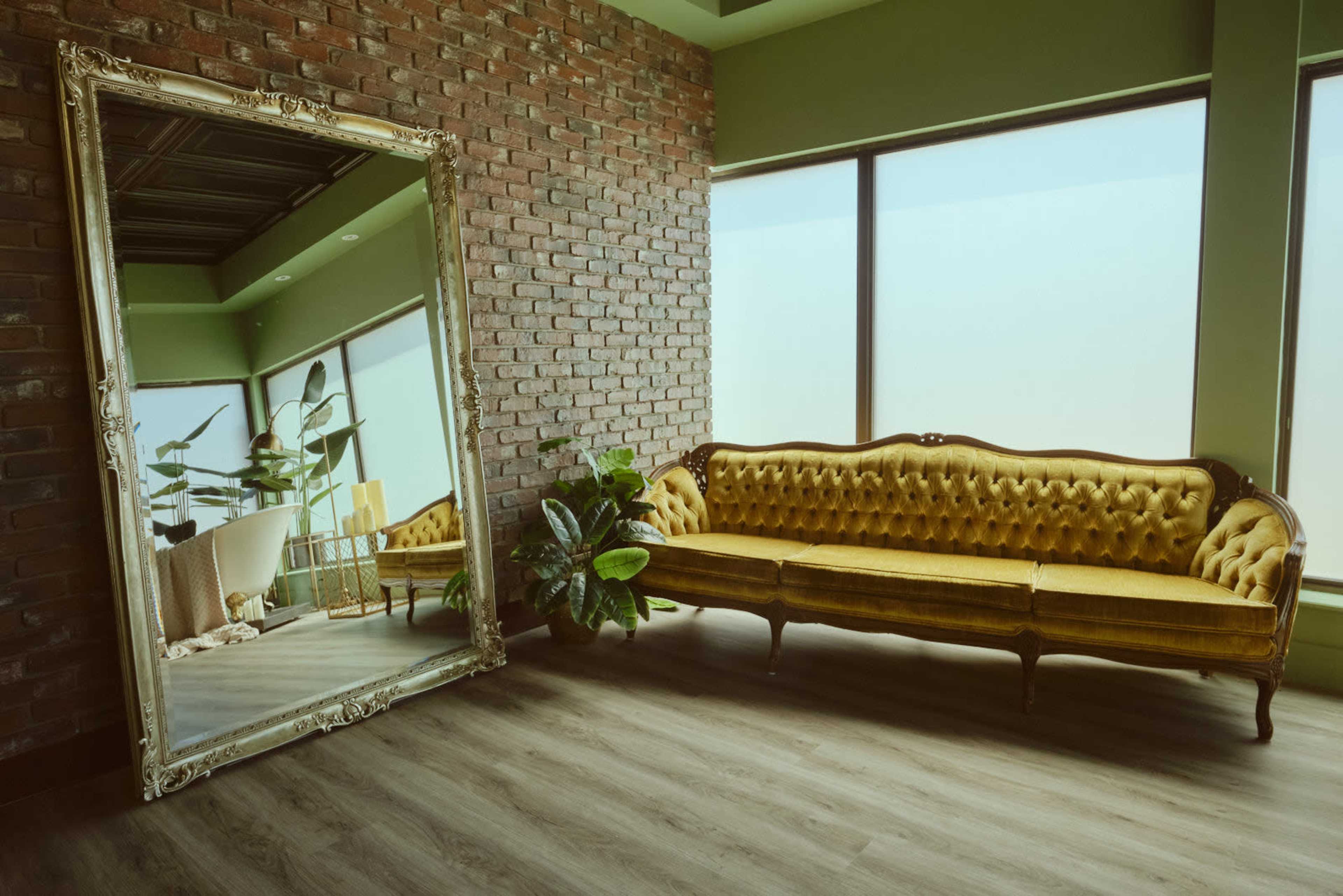 A vintage yellow sofa is positioned near a large mirrored wall in a room with exposed brick walls and large windows.