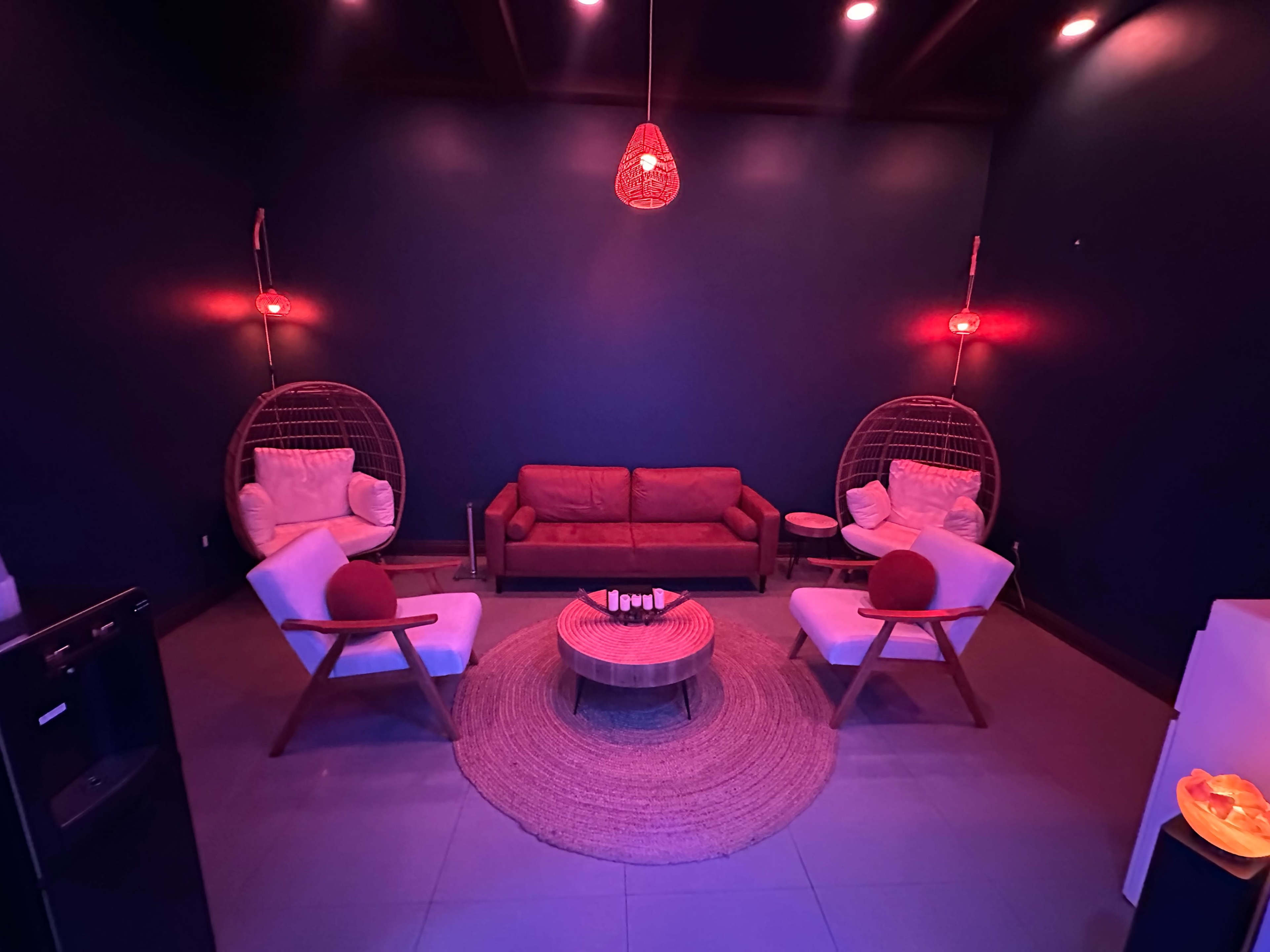 The image shows a dimly lit room with red accent lights, featuring a red sofa, two hanging chairs, and a round coffee table on a circular rug.