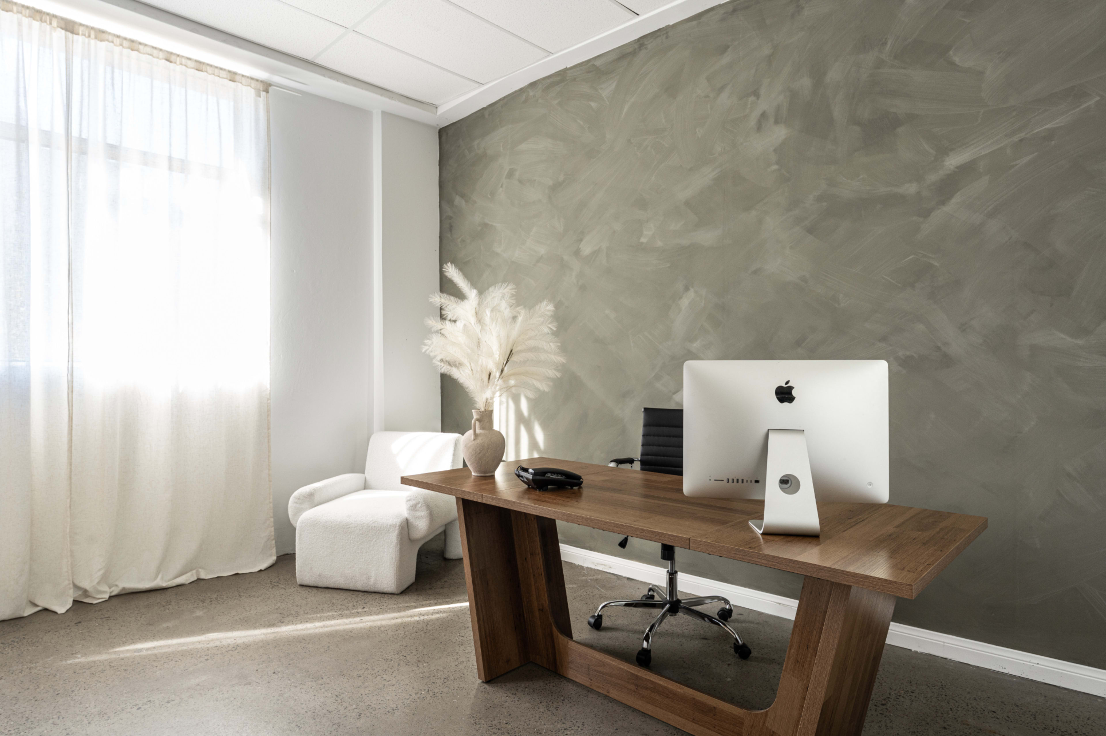 The image shows a modern office space featuring a wooden desk with an iMac, a black chair, a white armchair, and a large window with sheer curtains.