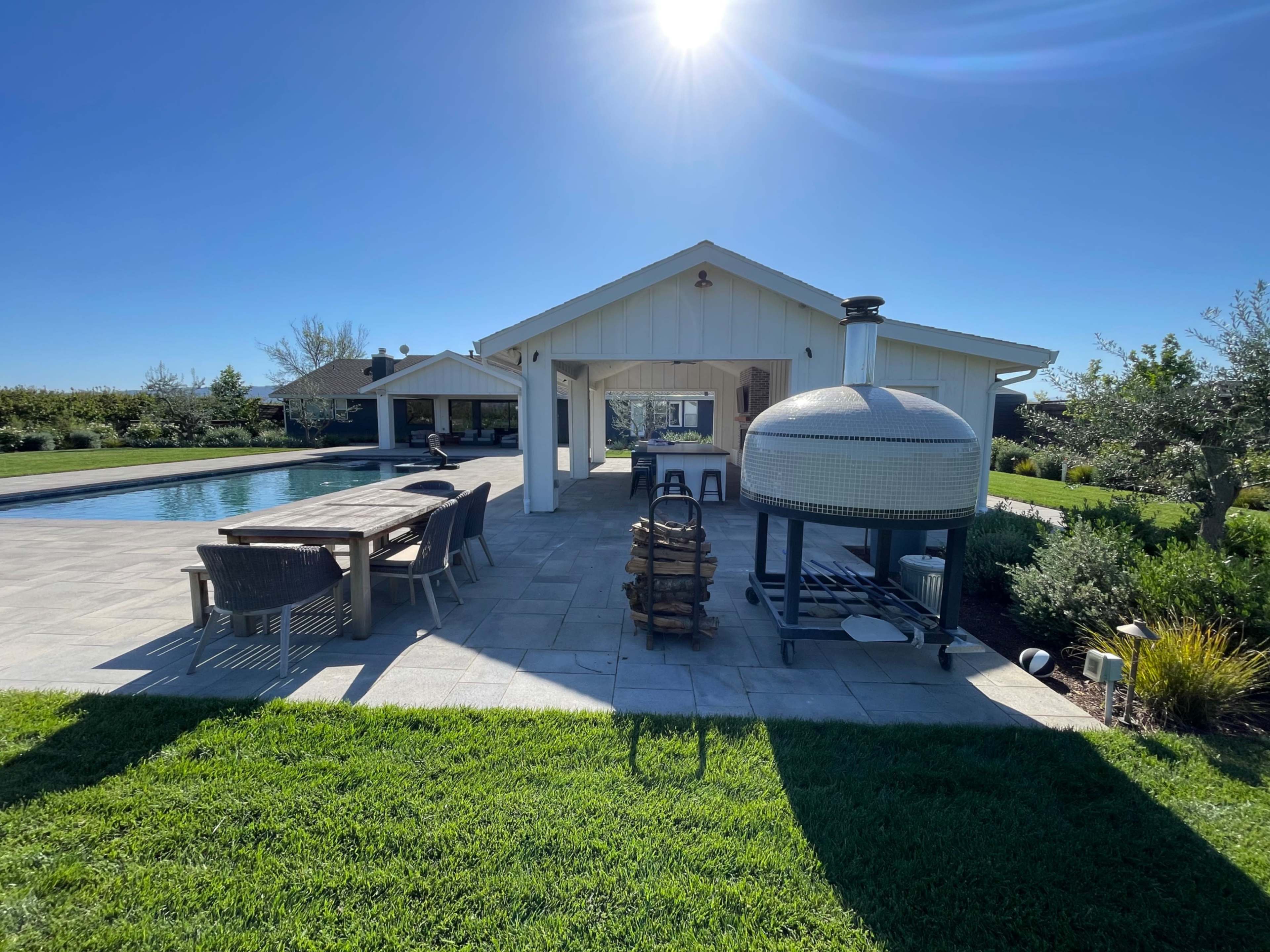 The image depicts a modern outdoor space featuring a swimming pool, a large dining table, and a circular grill near a white patio structure.
