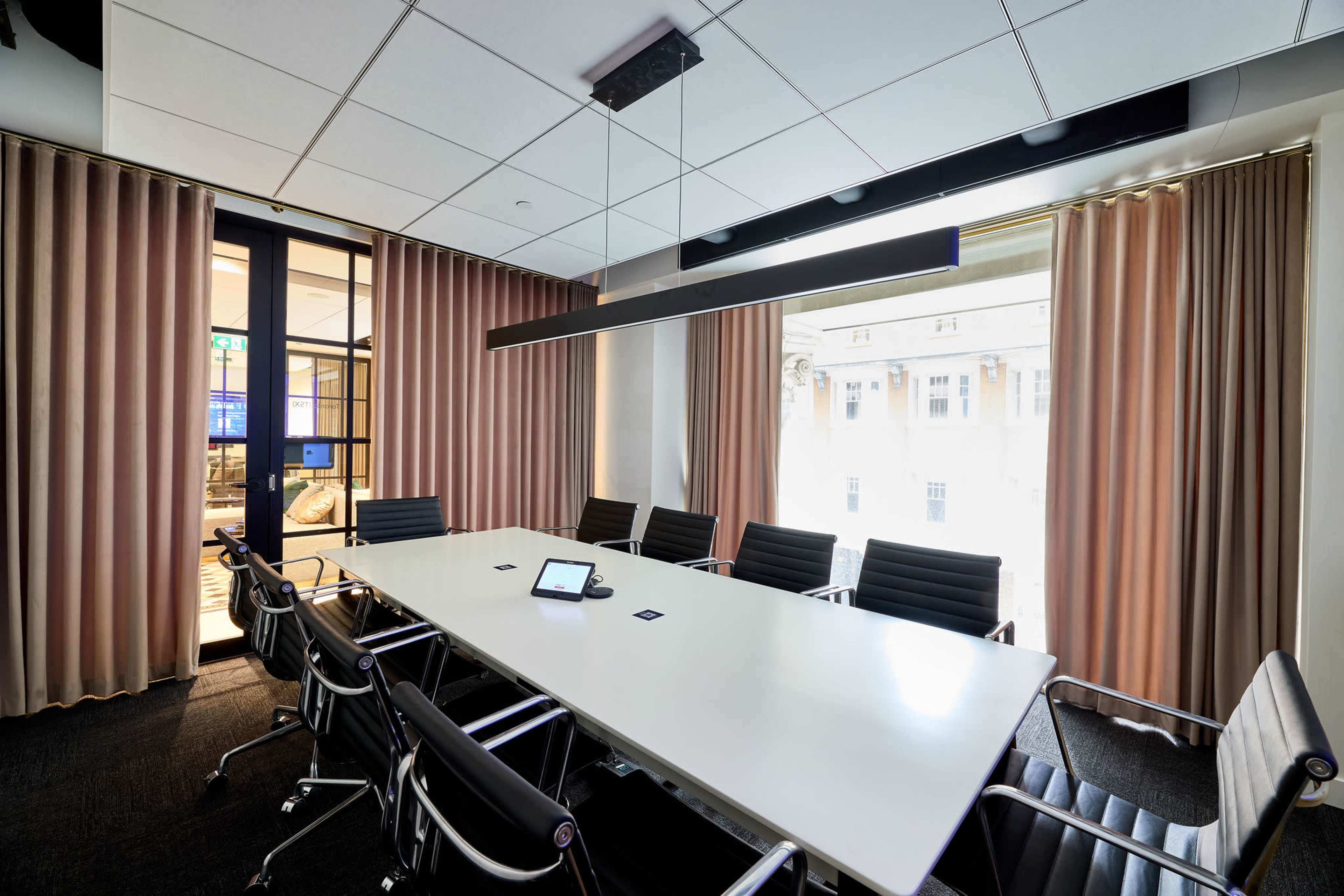 A large, modern conference room features a long white table surrounded by black office chairs, with large windows covered by curtains.