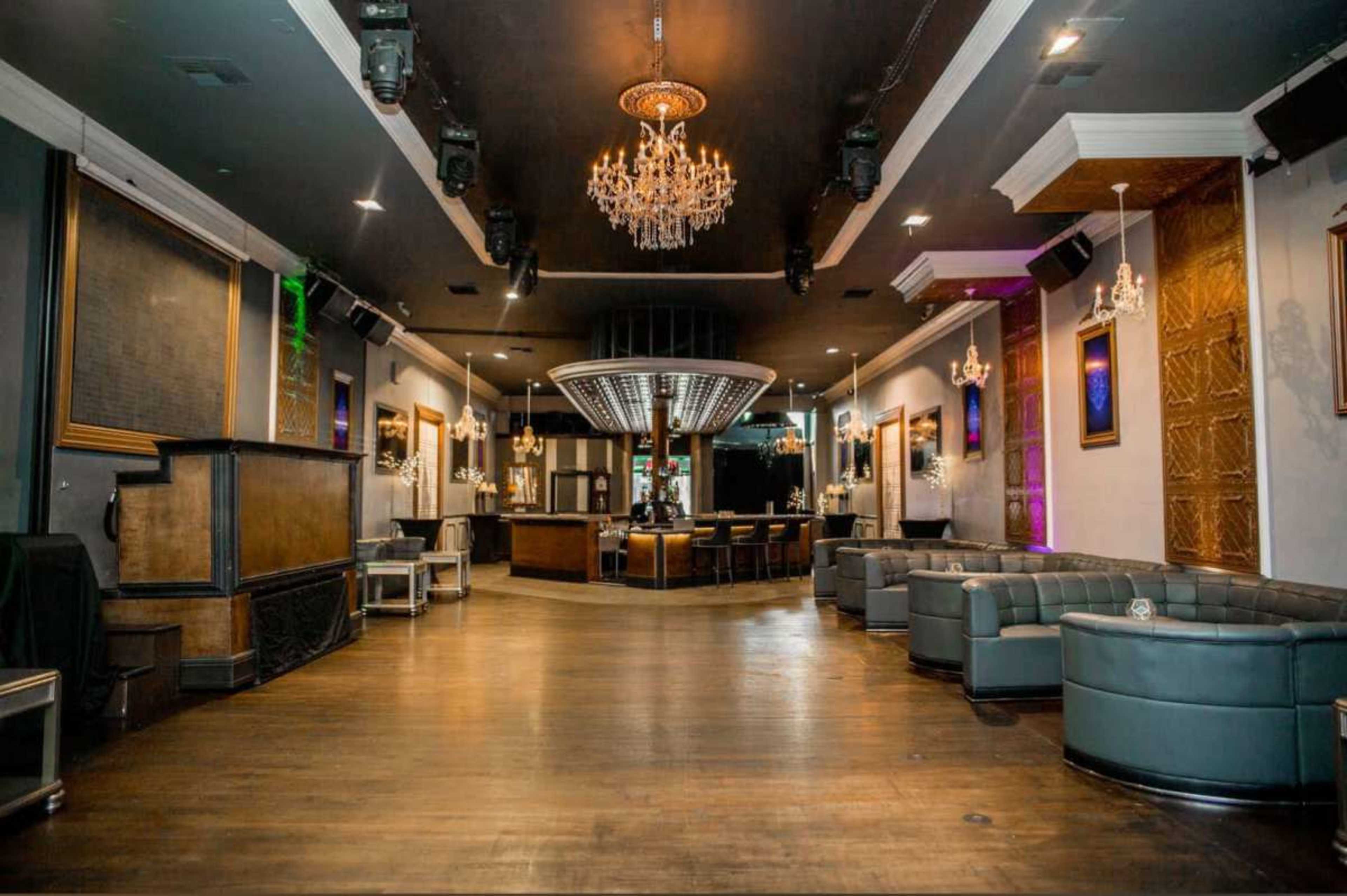 The image shows an empty upscale lounge area with dark walls, polished wooden floors, and a central bar surrounded by seating.