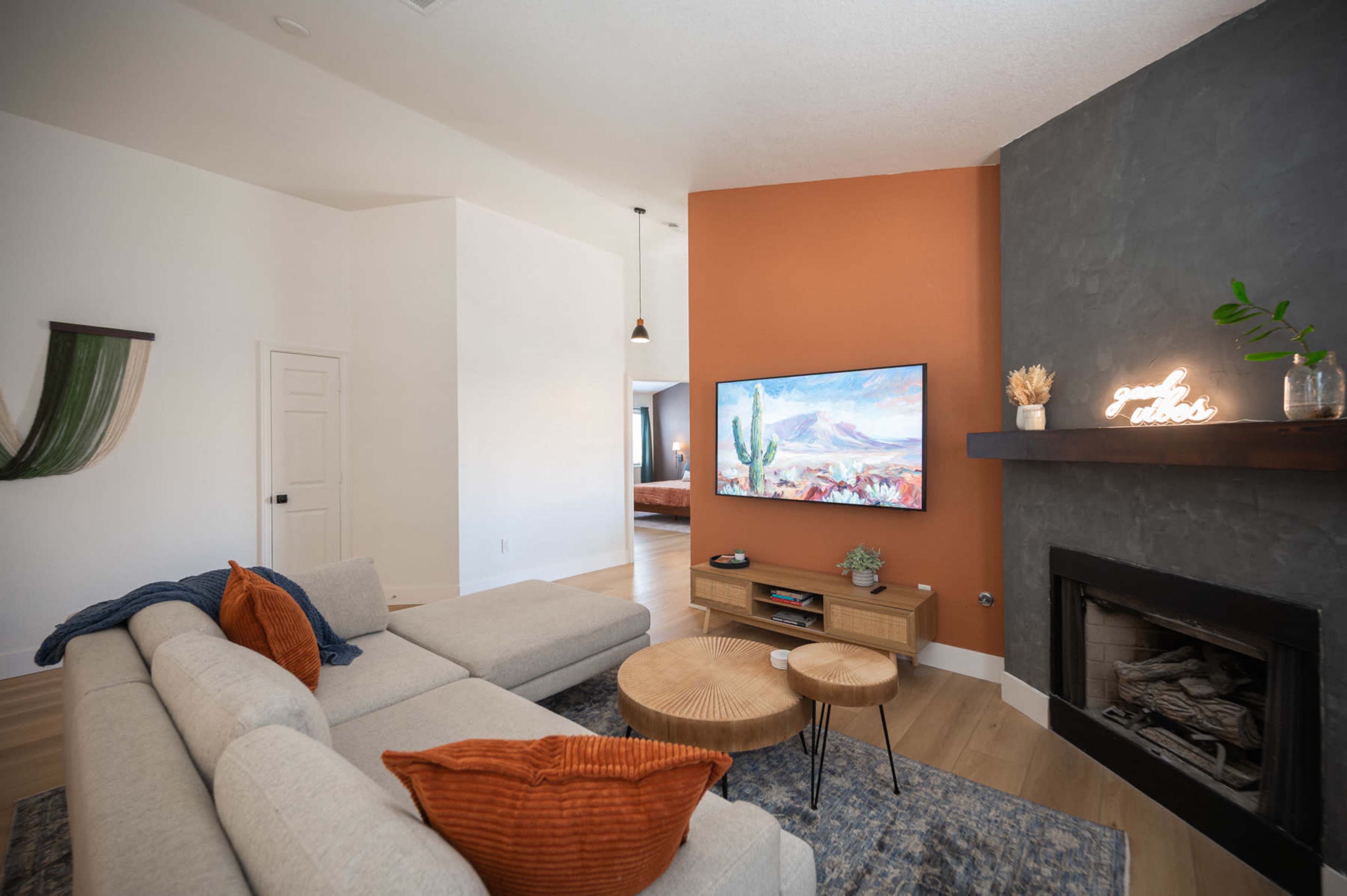 A modern living room features a sectional sofa with orange accent pillows, a wooden coffee table, and a wall-mounted television above a fireplace, set against an accent wall painted in terracotta.