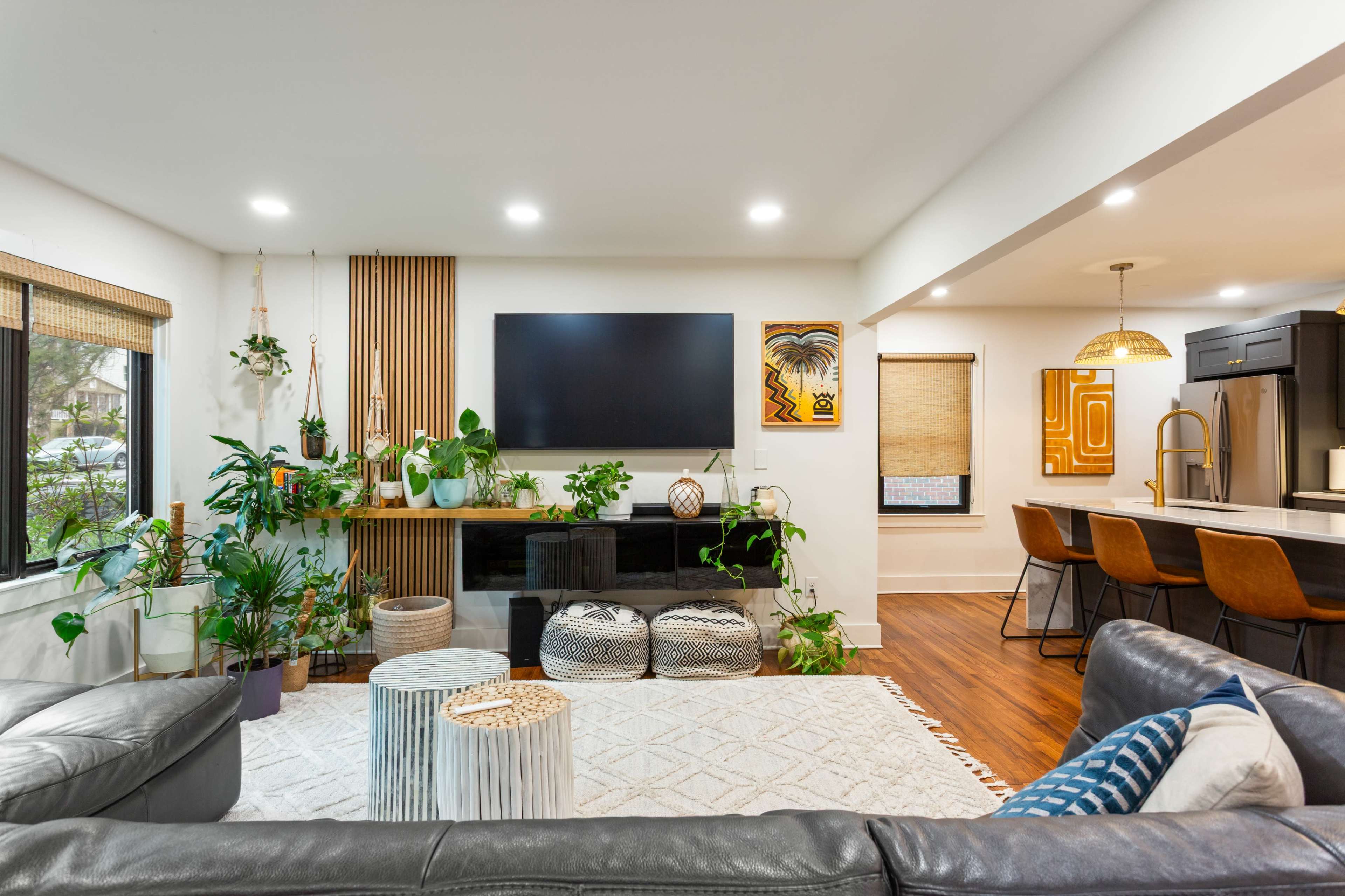 The room features a modern living area with a black TV mounted on the wall, surrounded by plants, a light rug, and a kitchen bar with four high-backed chairs.