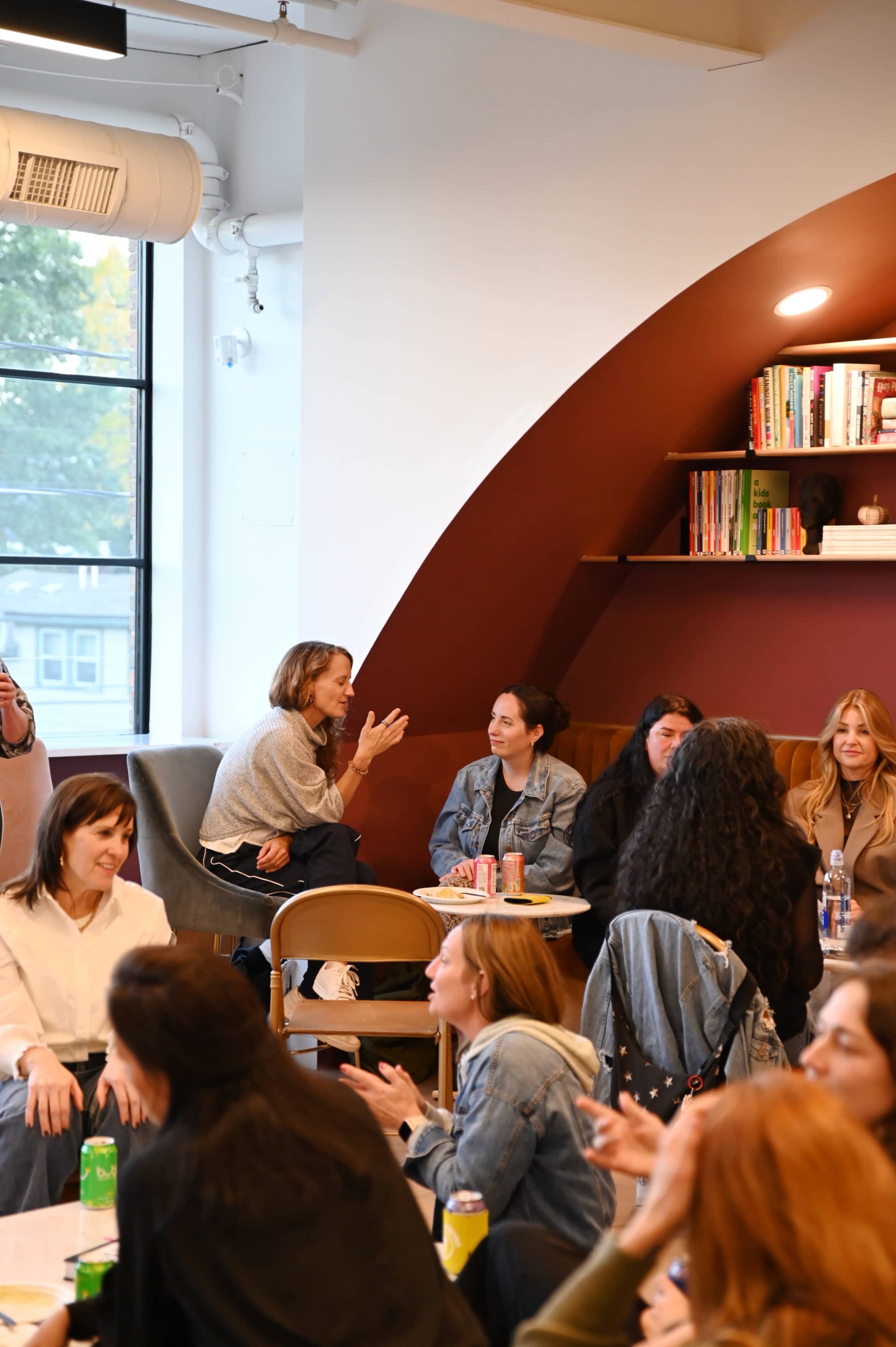 A group of women socialize in a cozy, modern café setting with bookshelves and large windows.