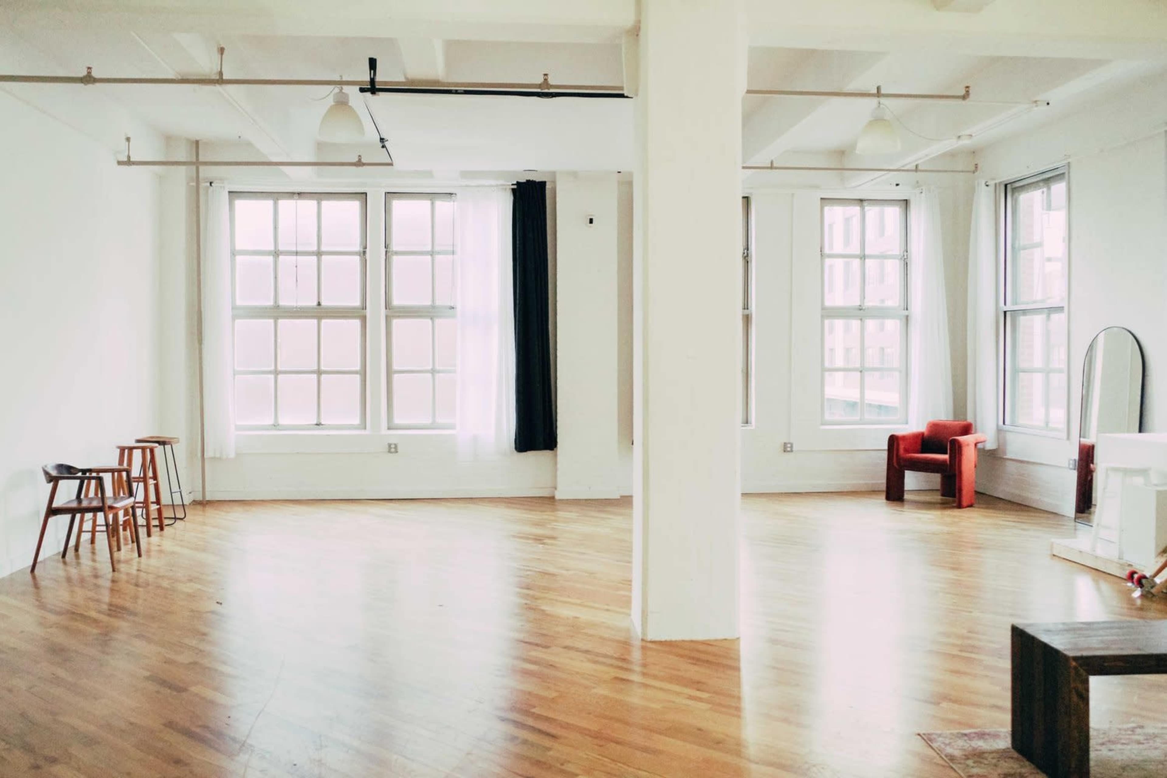 A spacious, empty room with hardwood floors, large windows, and minimal furniture including a red chair and a few wooden stools.