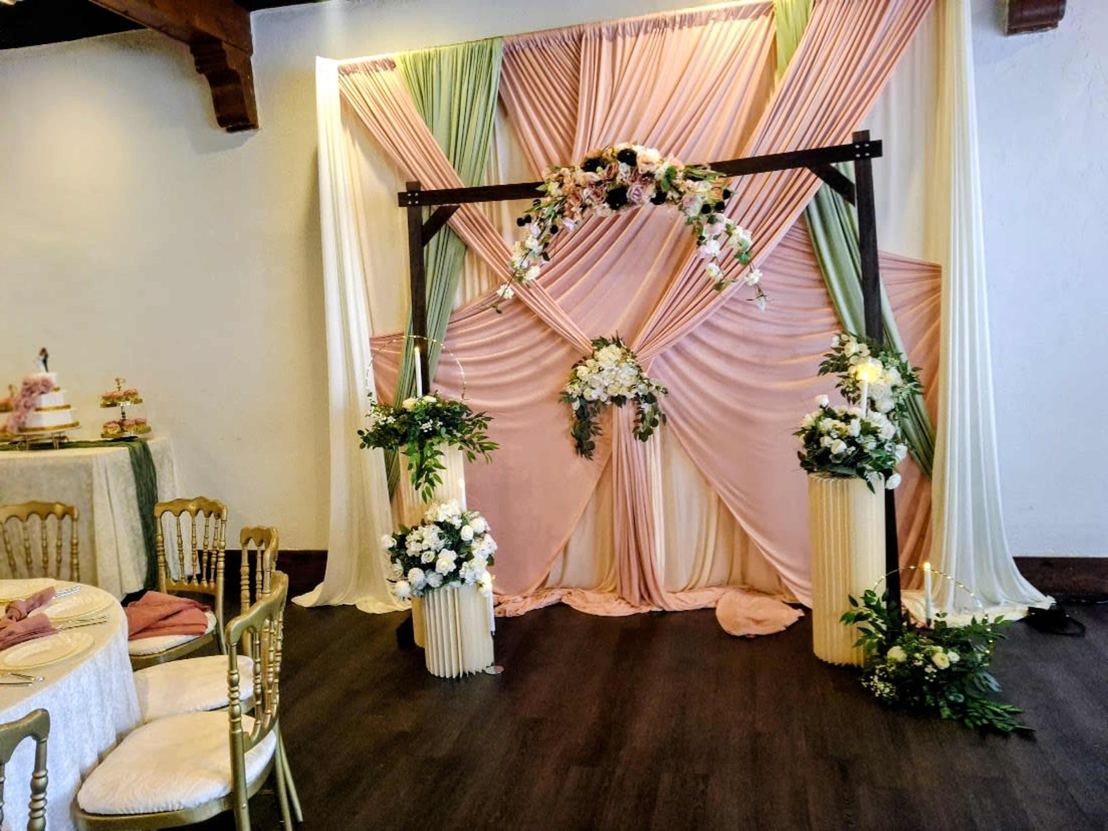 The image shows a decorated stage area with draped fabric in pastel colors, floral arrangements, and decorative columns, set against a neutral wall.