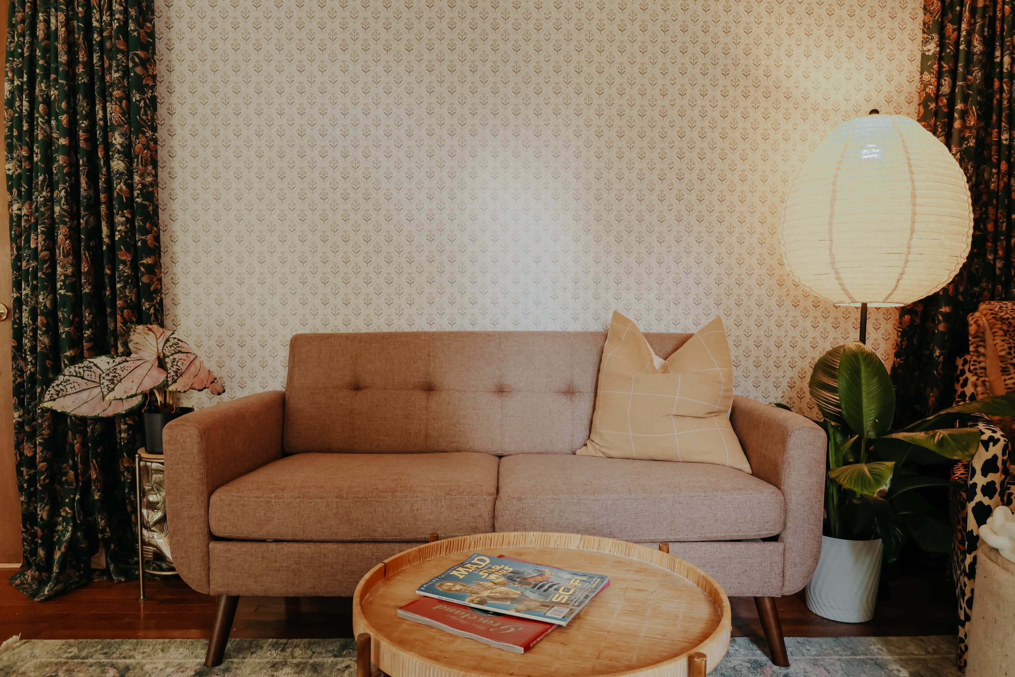 A cozy living room features a light brown sofa with a decorative pillow, a round wooden coffee table, and a tall lamp beside a potted plant and patterned curtains.