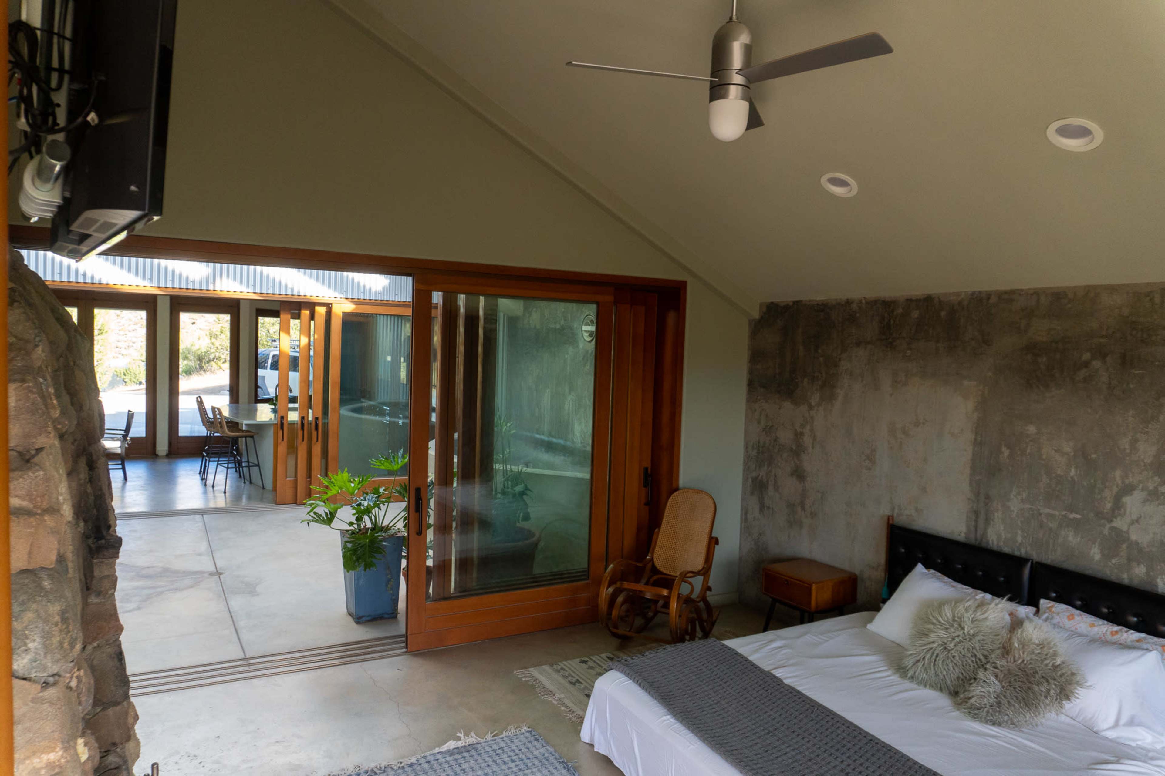 A bedroom with a bed and a wooden rocking chair, leading to a glass sliding door that opens to a bright living area.