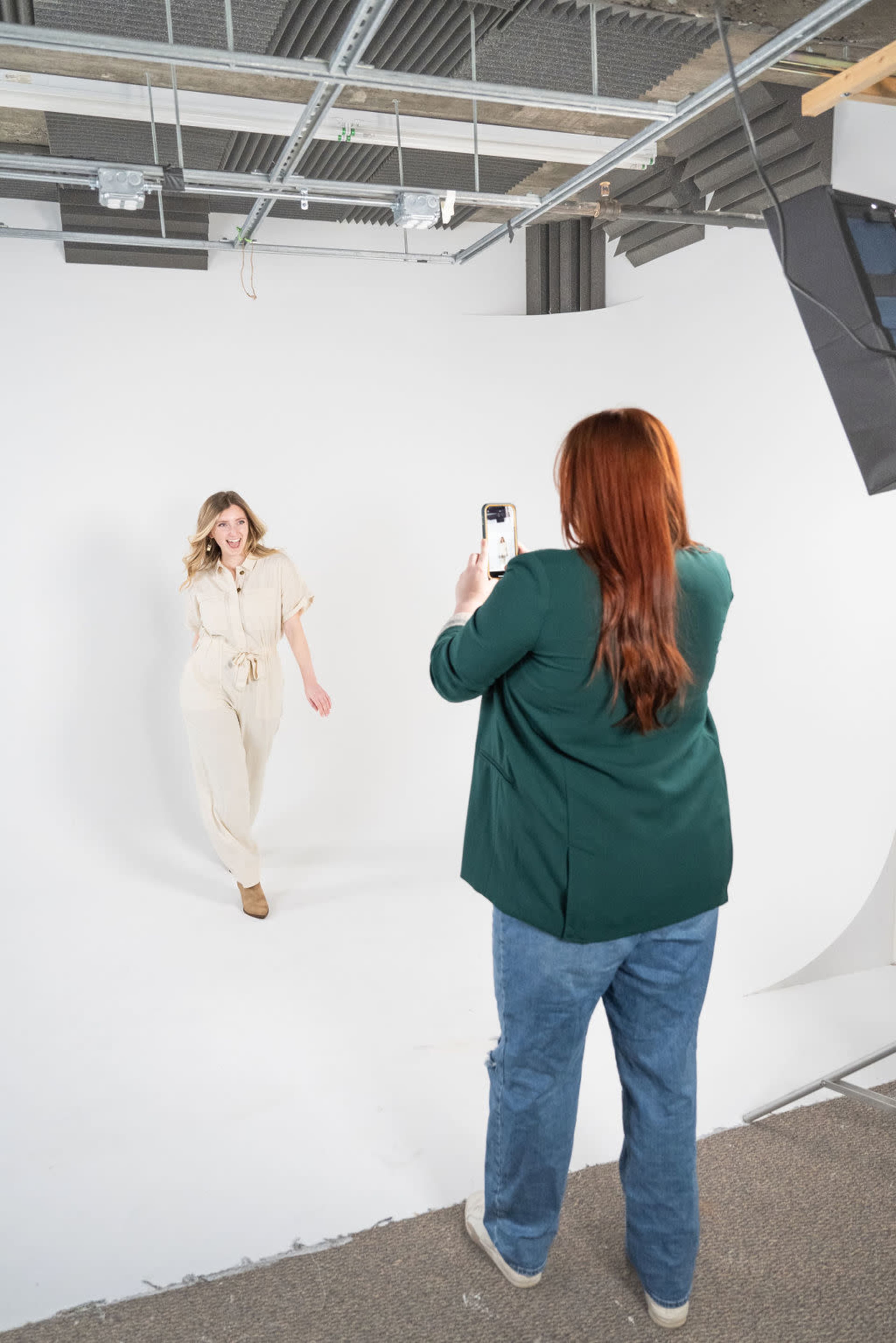 A woman takes a photo of another woman posing in a studio with a white backdrop.