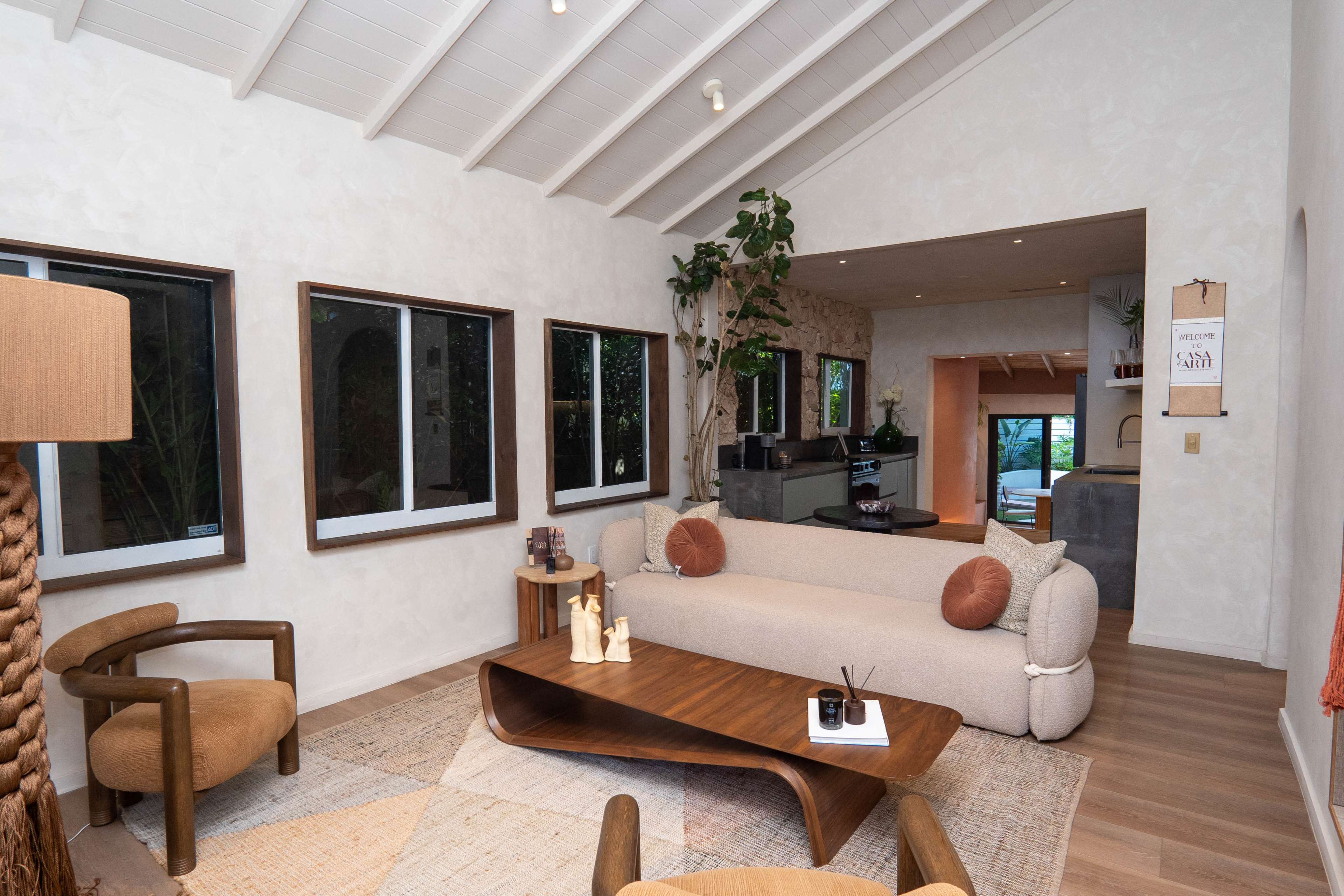 The image shows a modern living room with a curved, light-colored sofa, a wooden coffee table, and large windows that let in natural light.