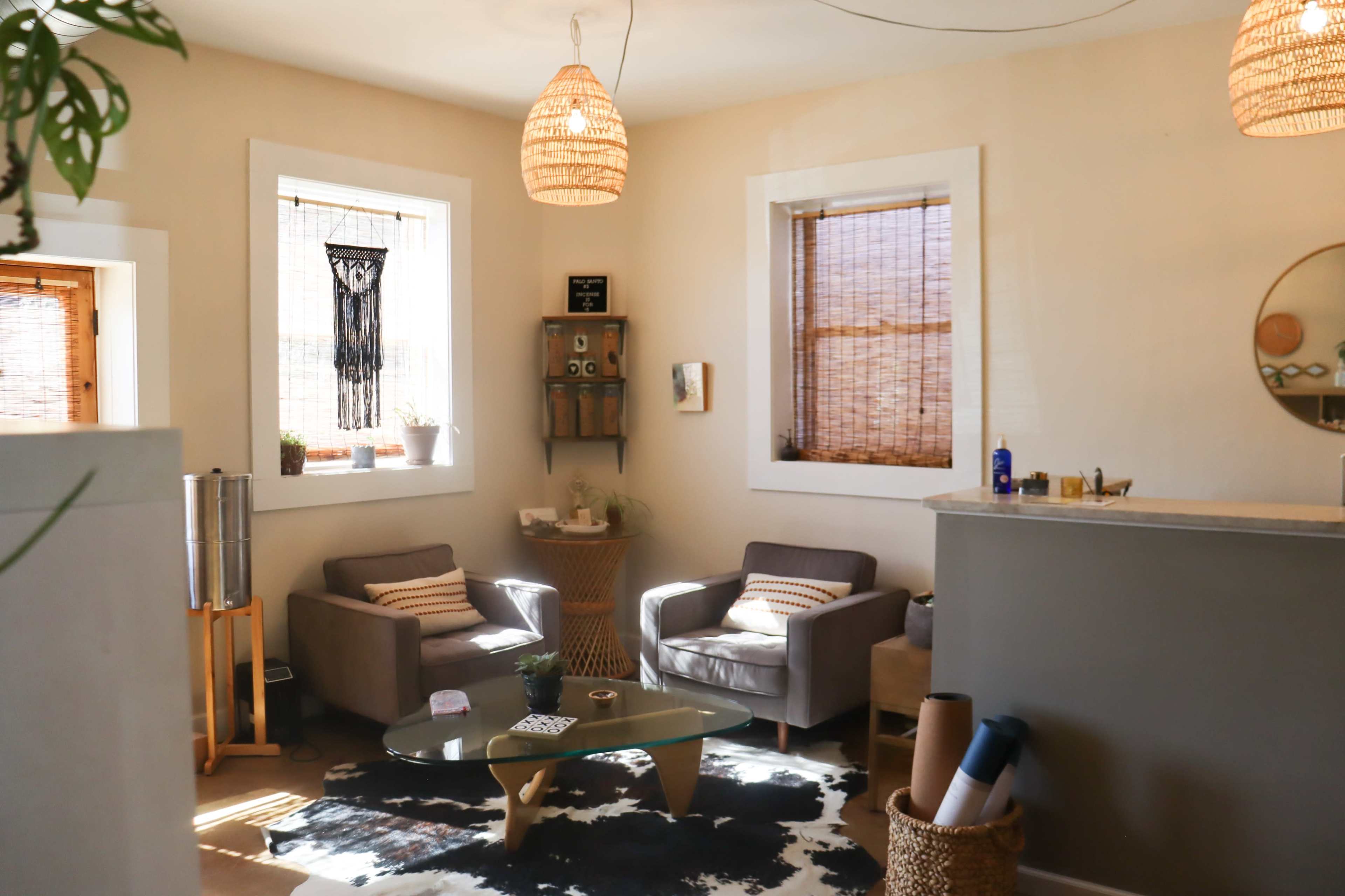 A cozy room features two gray armchairs, a glass coffee table, and natural decor elements, illuminated by pendant lights.
