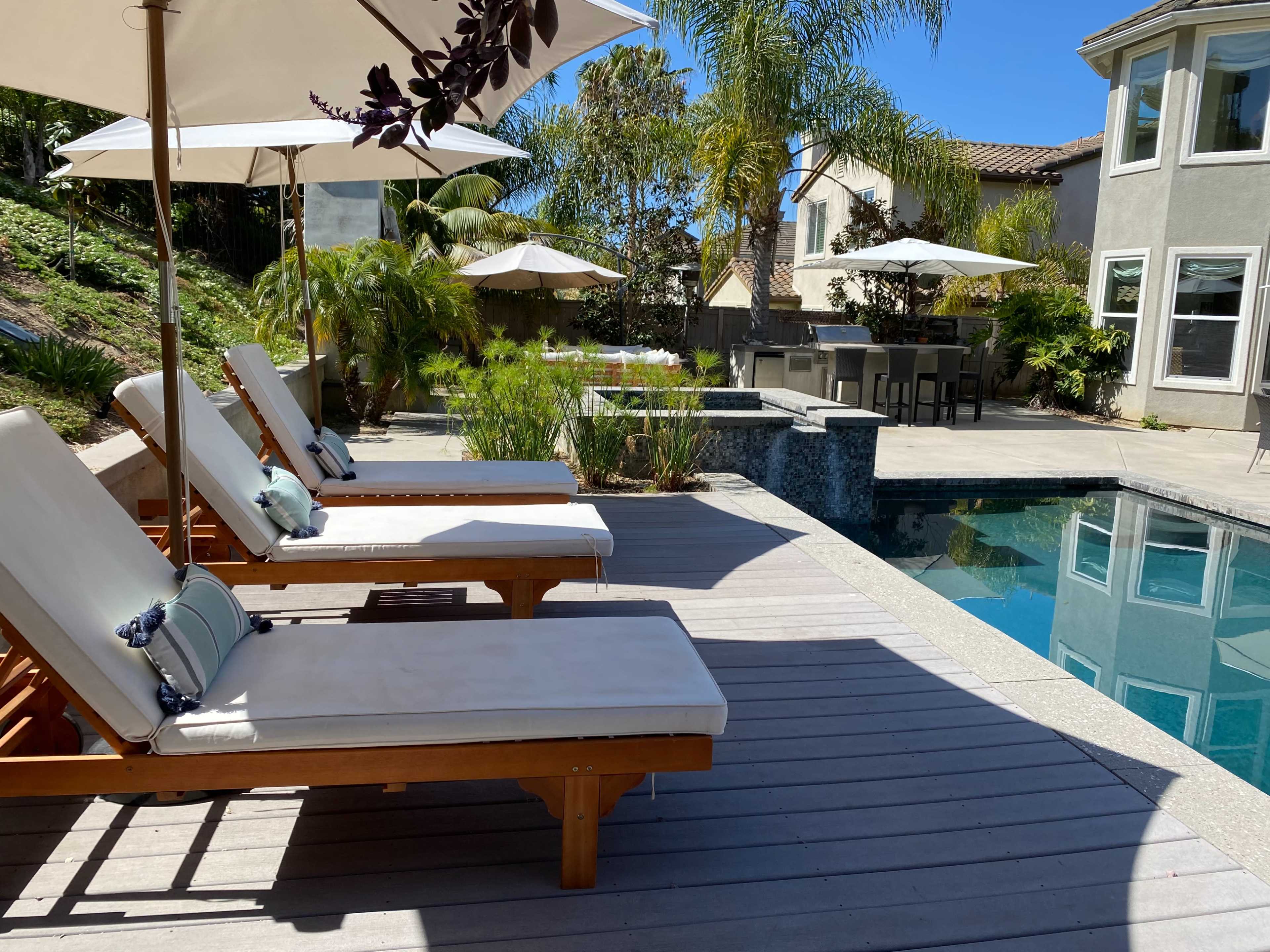 A poolside area features wooden lounge chairs and umbrellas, surrounded by landscaped greenery and a clear blue swimming pool.
