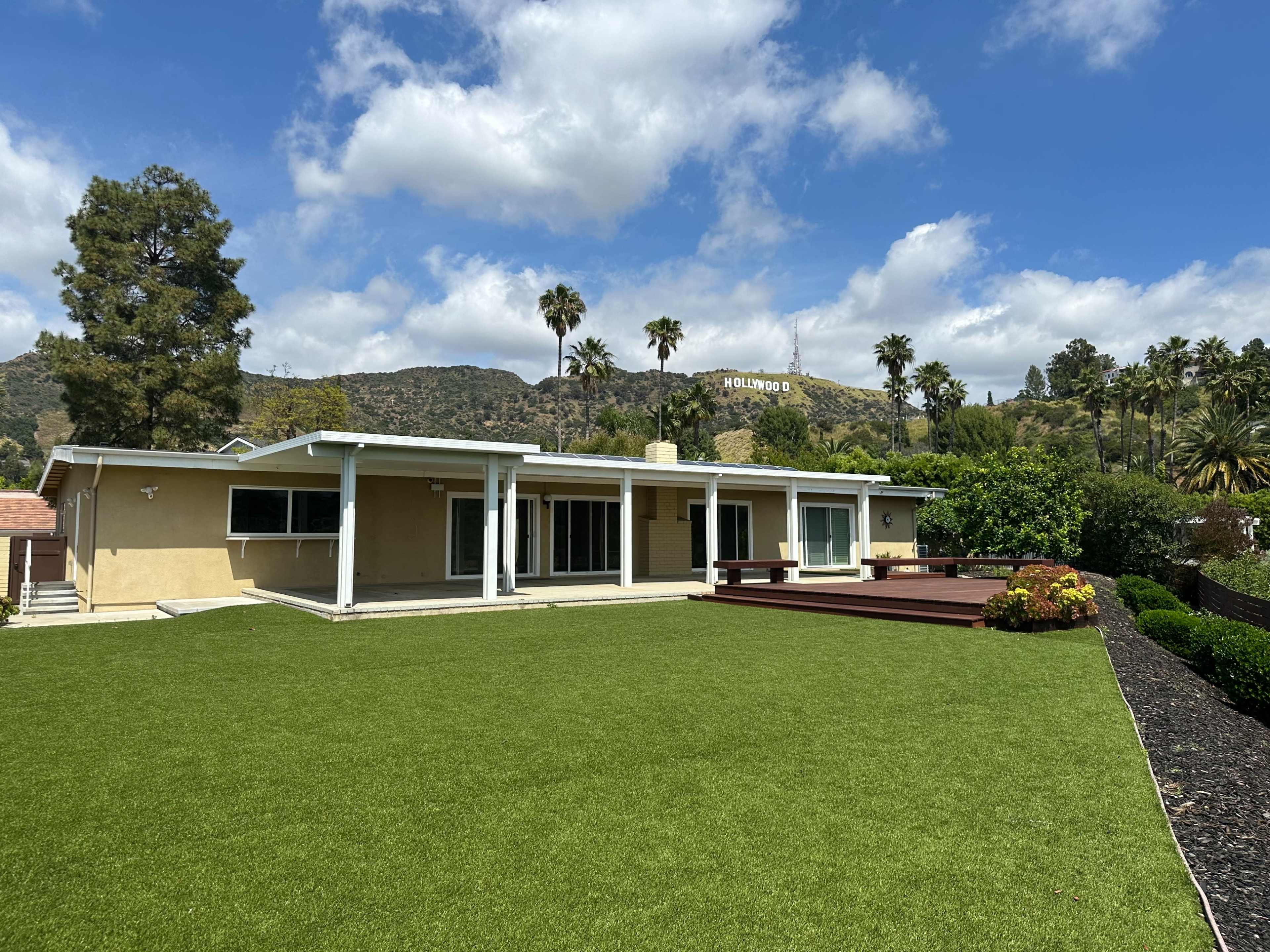 A single-story house with a large lawn and a wooden deck is situated against a hillside with palm trees, featuring the Hollywood sign in the background.
