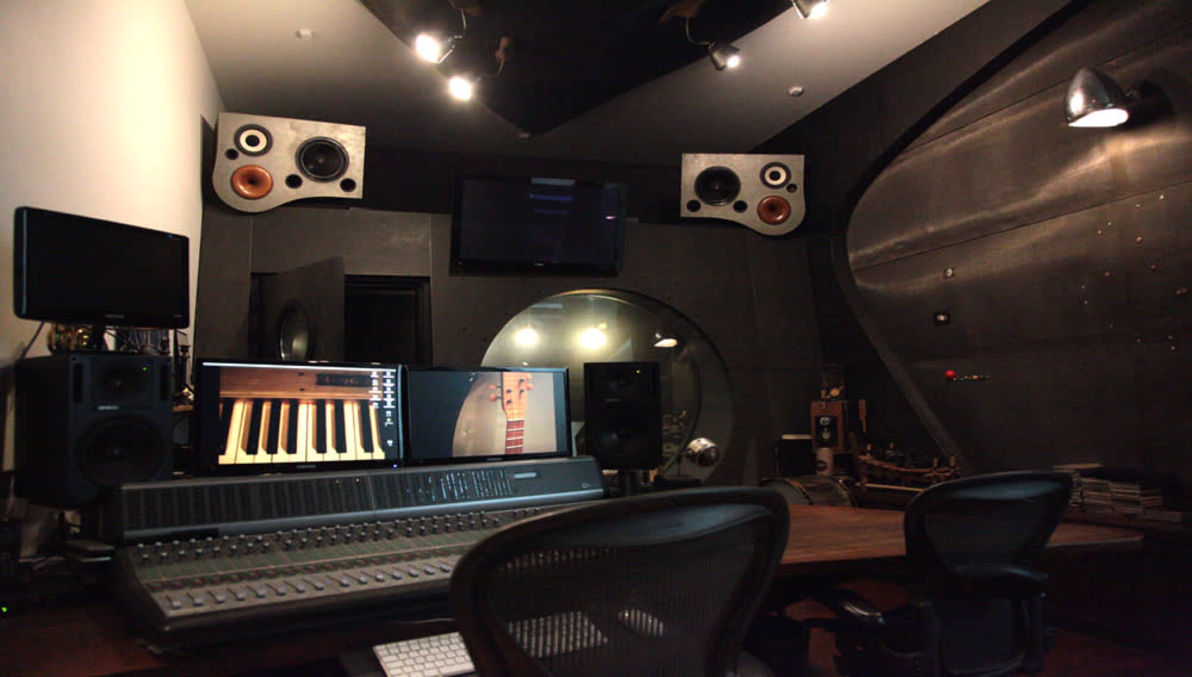 A professional sound recording studio with a mixing console, multiple speakers, and a large circular window.