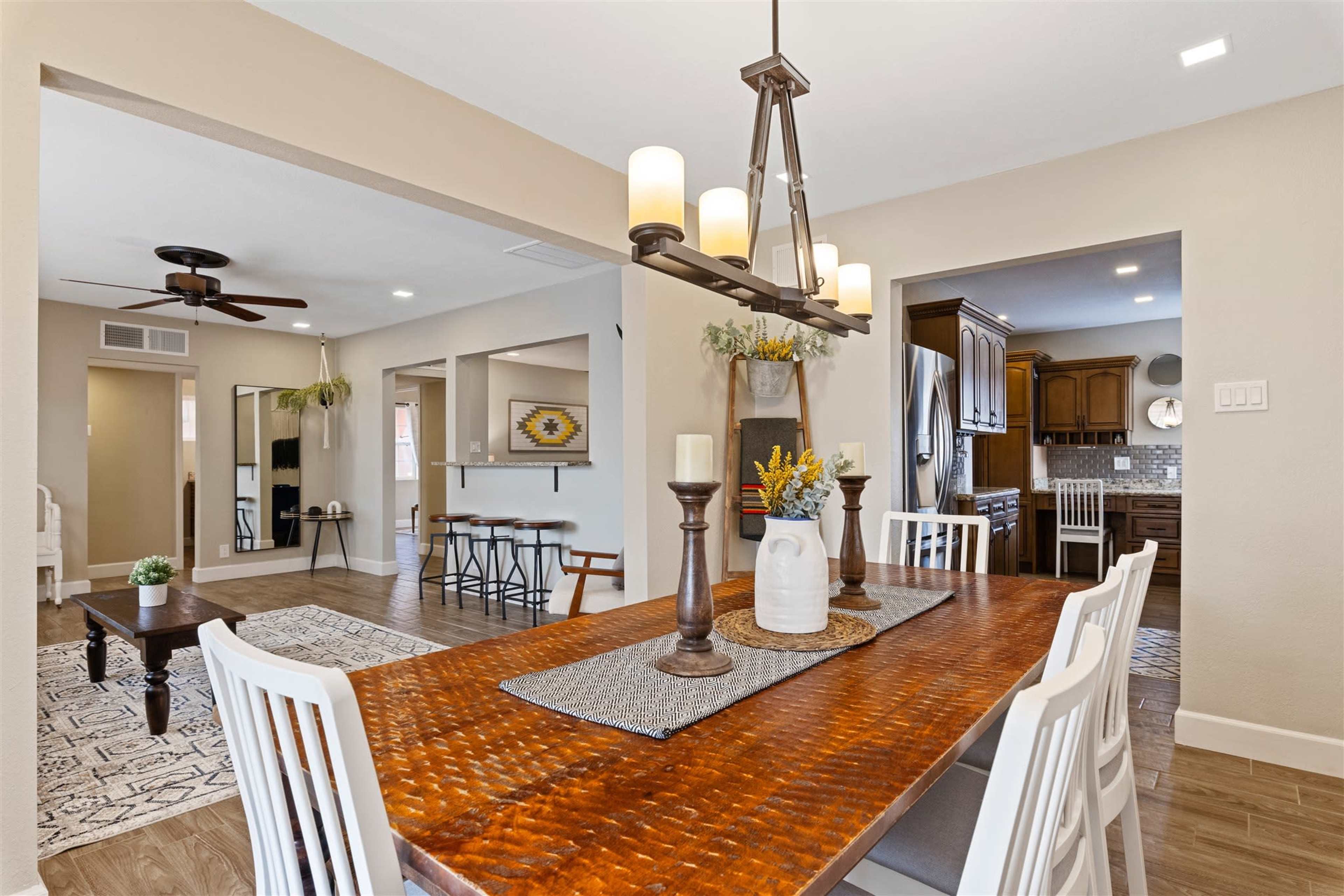 A dining area with a wooden table surrounded by white chairs, leading to an open-concept living space with a ceiling fan and kitchen area in the background.