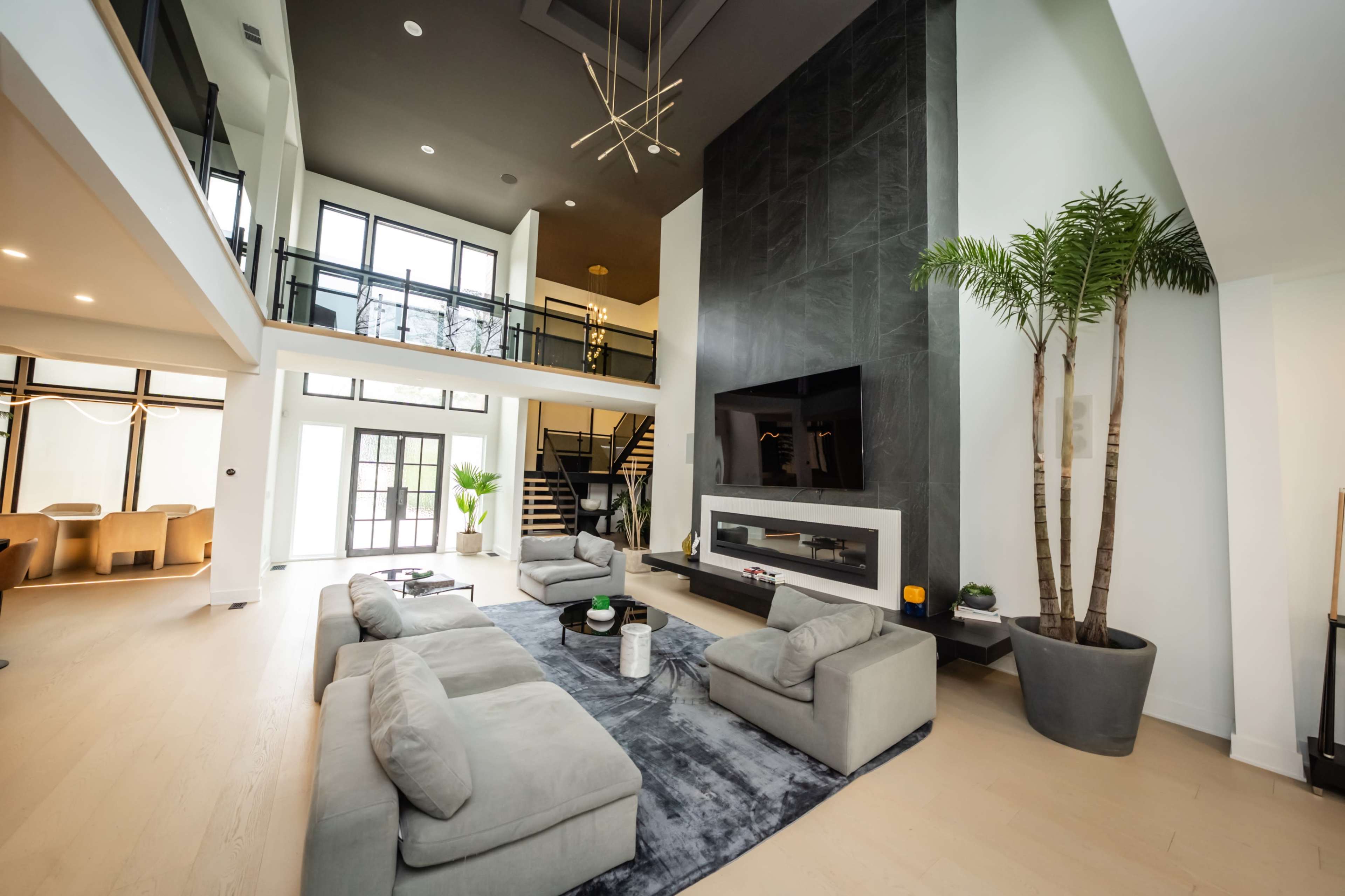 The image shows a spacious modern living room with large windows, a high ceiling, a black stone fireplace, and a sectional sofa arranged around a coffee table.
