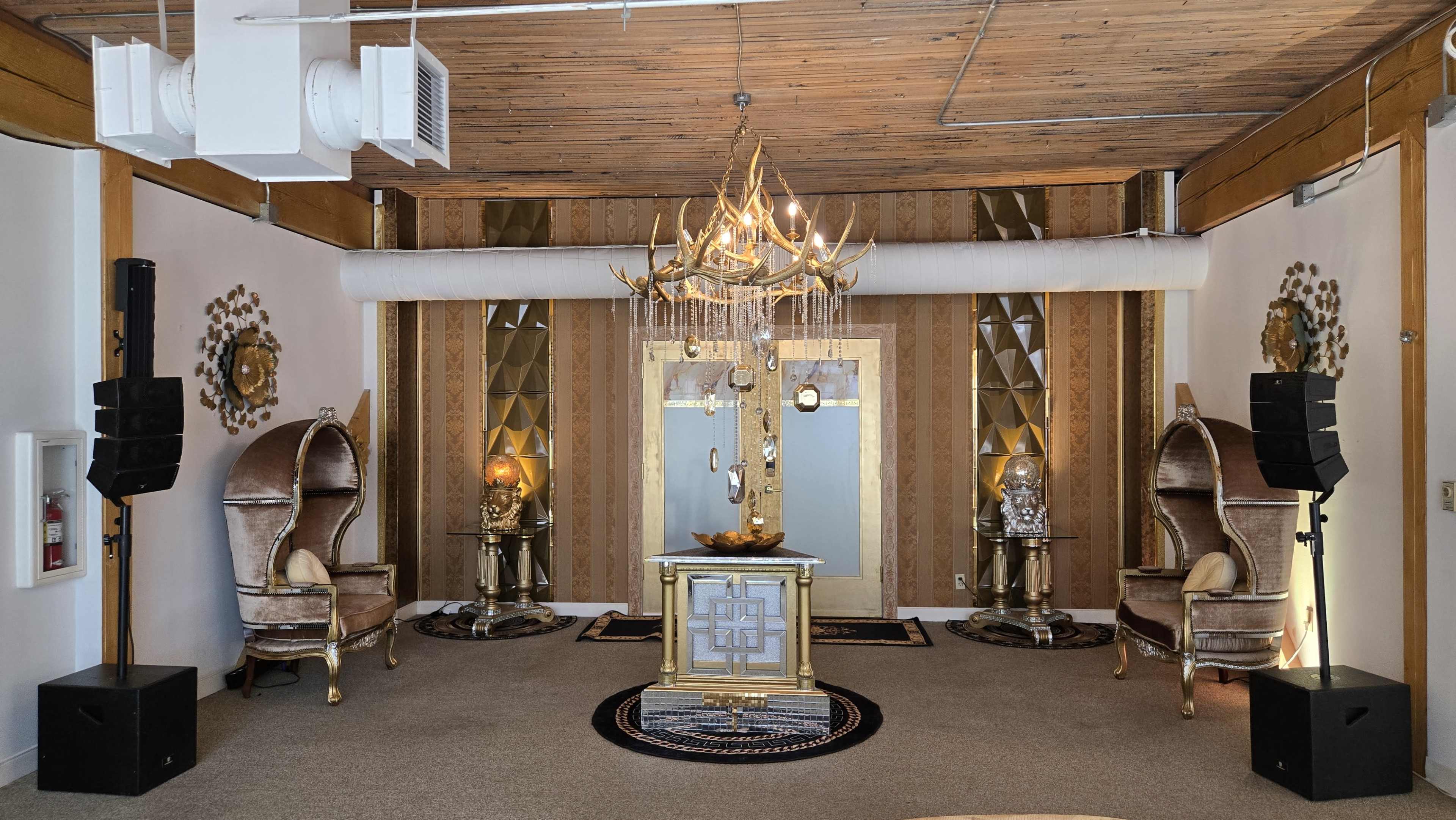 The image shows an elegantly decorated room featuring ornate furniture, a central decorative table with a chandelier above, and speaker setups on either side.