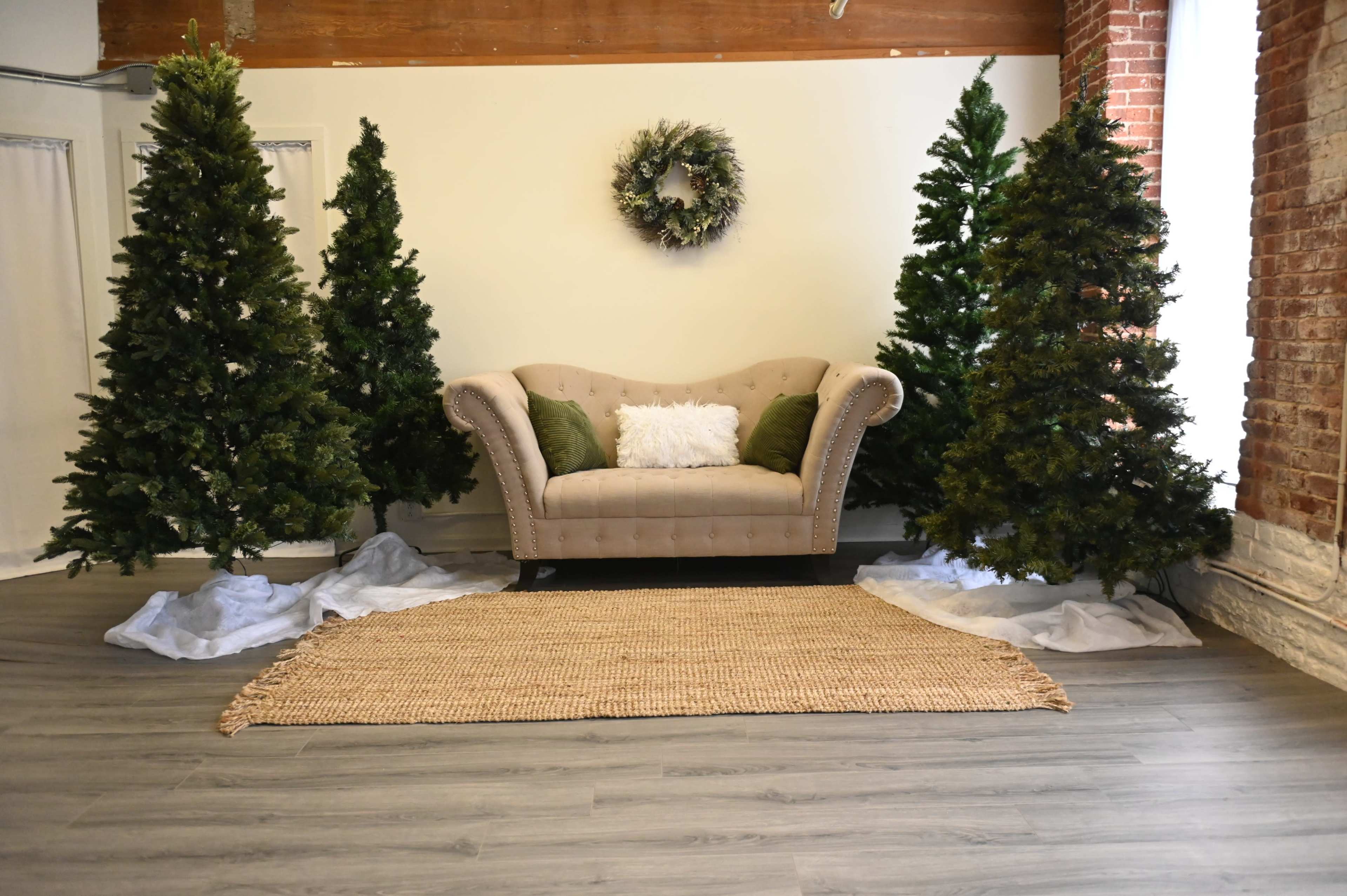 A tufted beige couch is positioned in front of two decorated Christmas trees, with a woven rug on the floor and a wreath hanging on the wall above the couch.