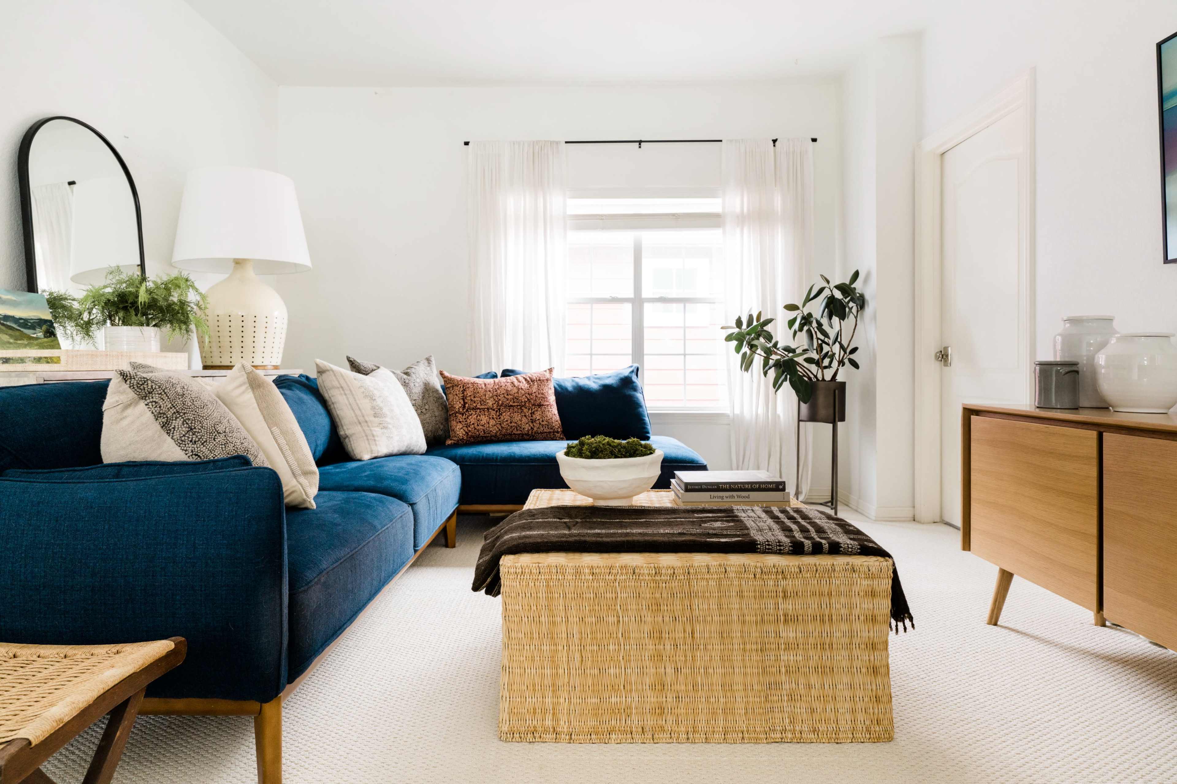 The image shows a modern living room with a blue sectional sofa, a round coffee table made of woven material, and a wooden sideboard against the wall.