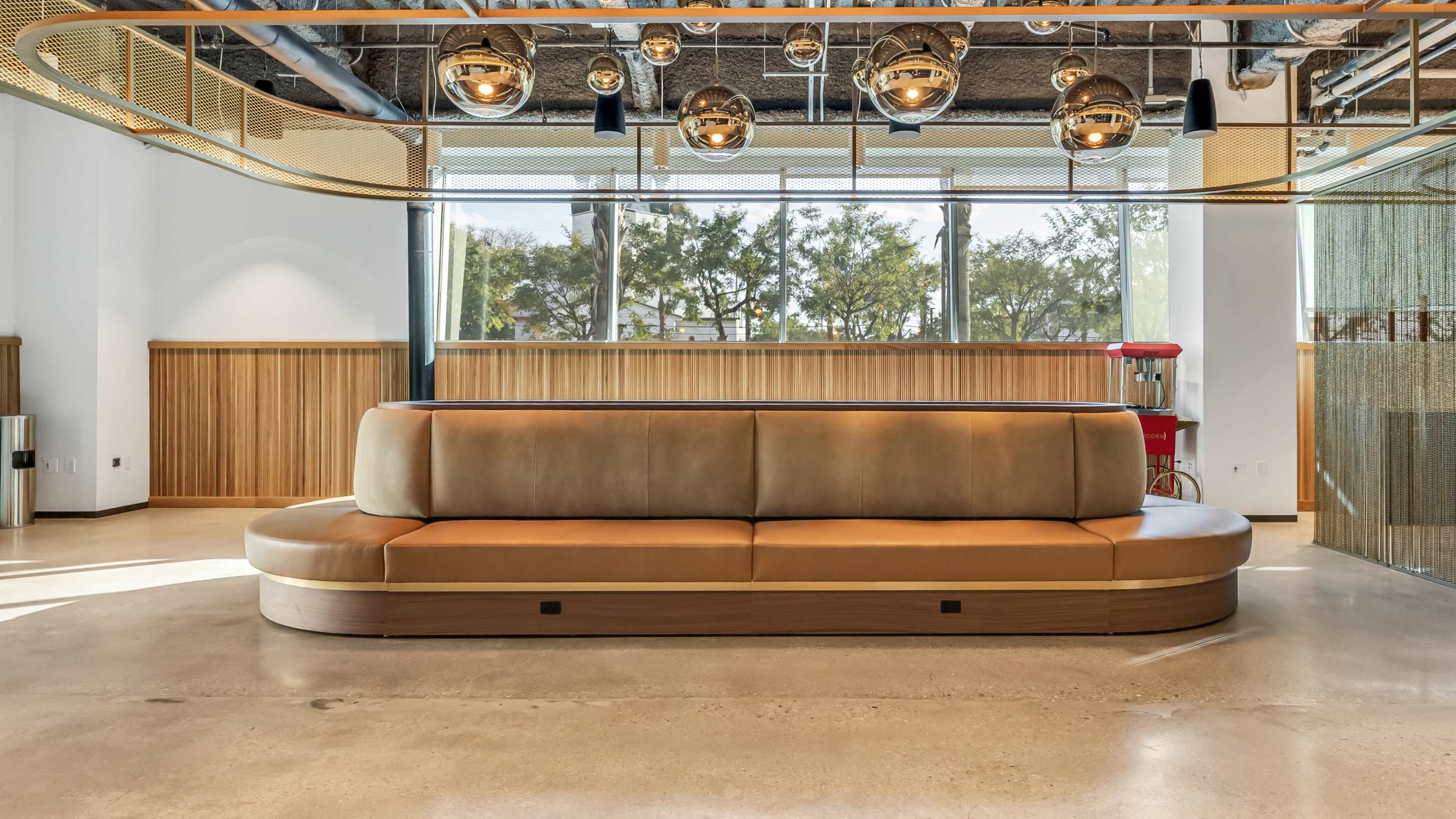 A modern, curved brown leather sofa positioned under spherical light fixtures in a well-lit space with large windows and wooden accents.