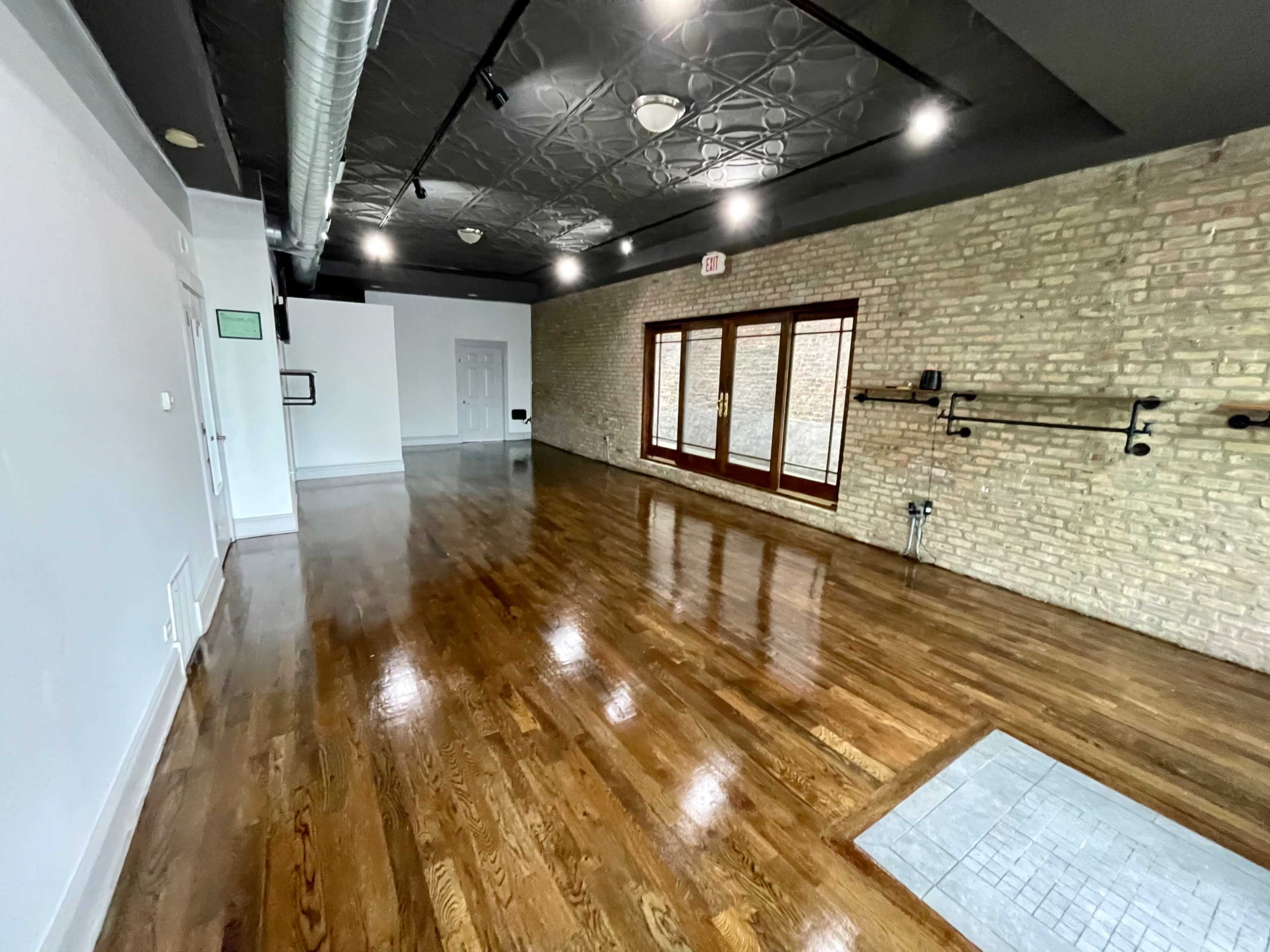 A spacious, empty room with wooden floors, exposed brick walls, and large windows allowing natural light to enter.