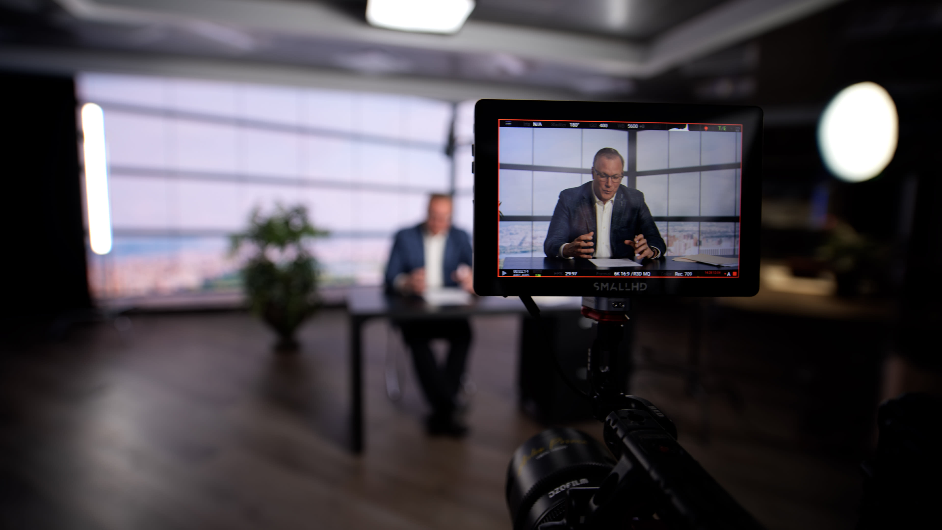 A man is seated at a table in an office setting, being filmed for a video presentation.