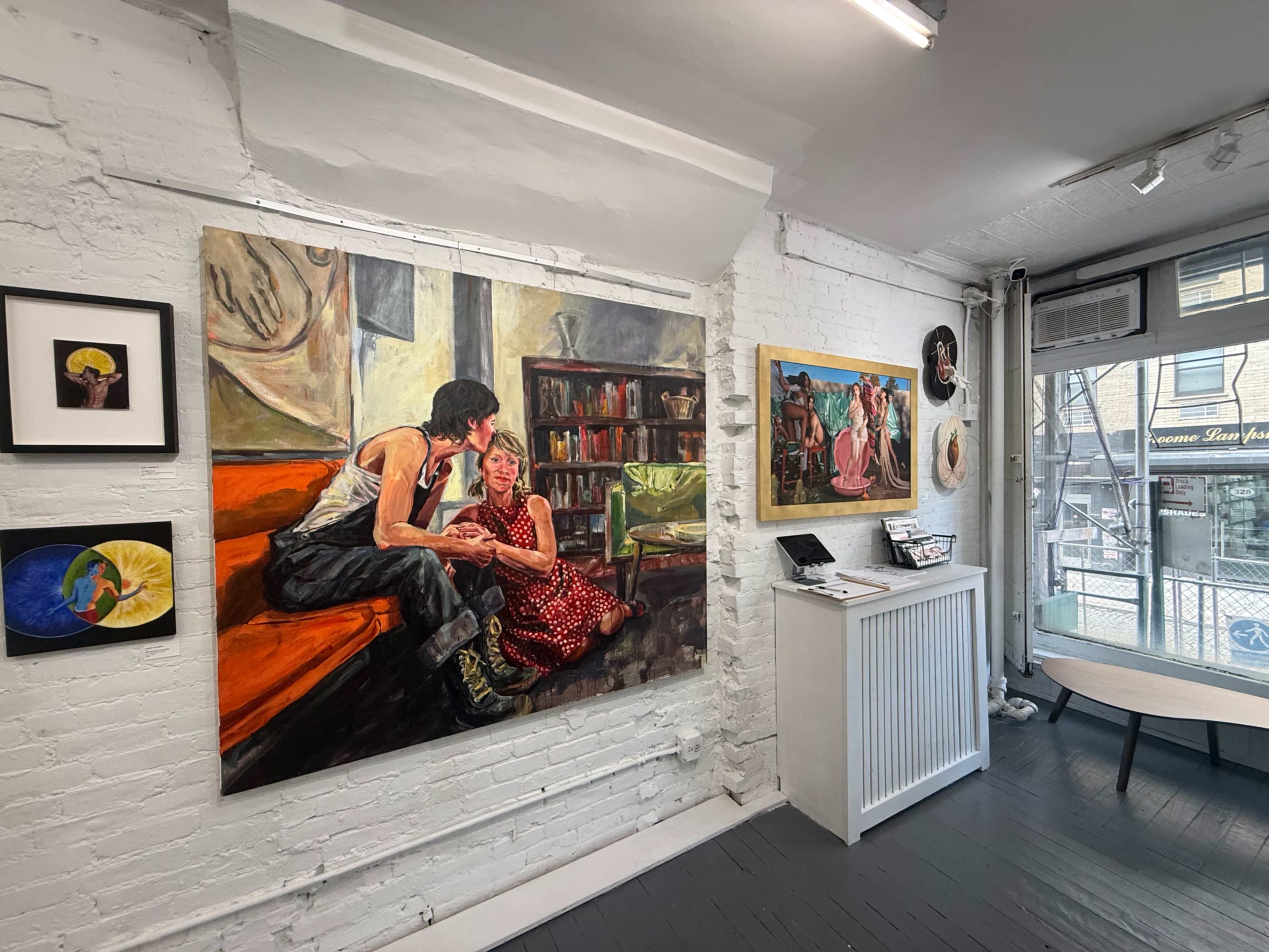 The image shows an art gallery featuring several paintings on white-brick walls, with a colorful scene of two people conversing amidst shelves of books.