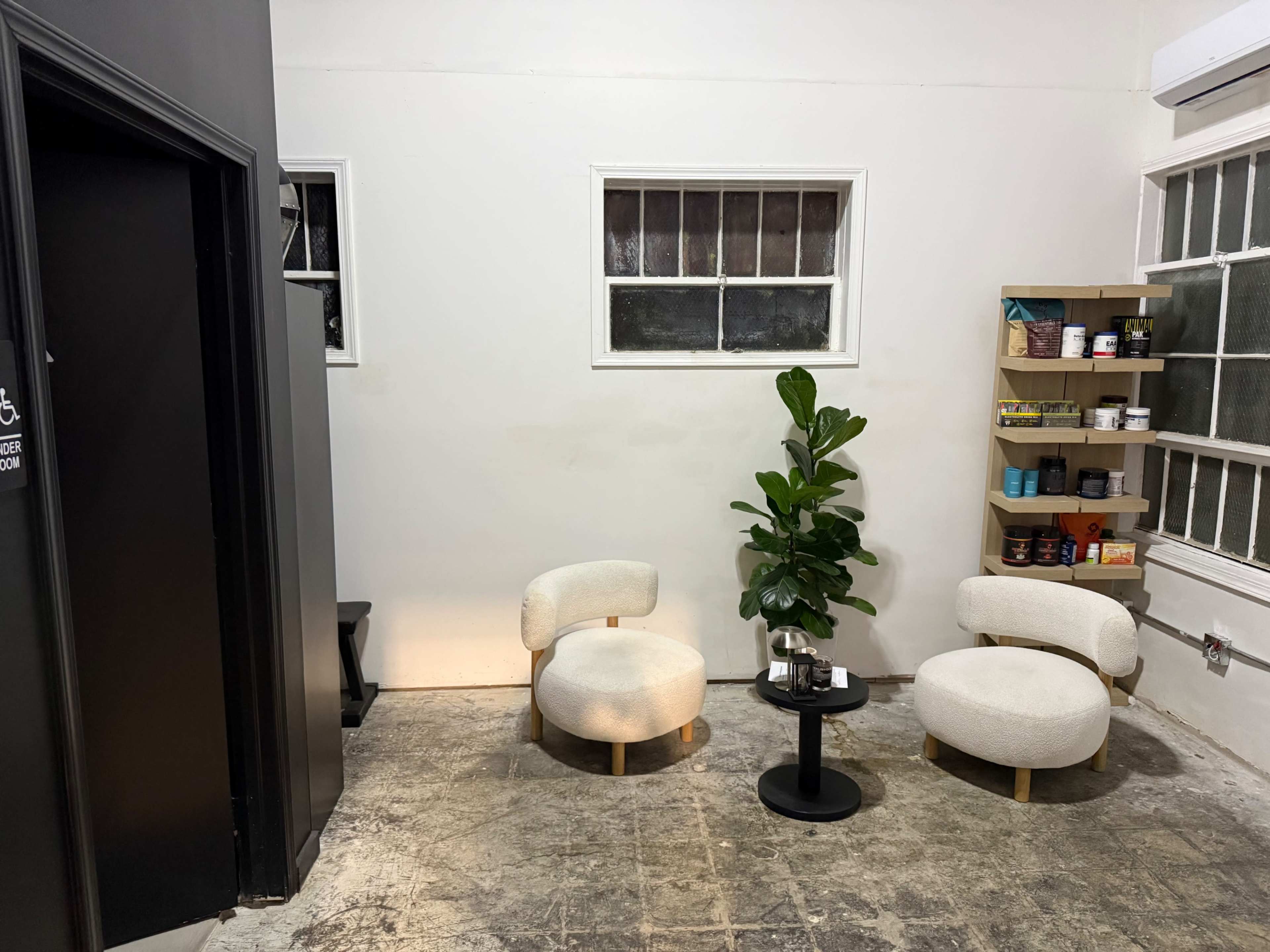 The image shows a small, minimalist lounge area with two round chairs, a small black table, a potted plant, and a shelf displaying various containers on the wall.