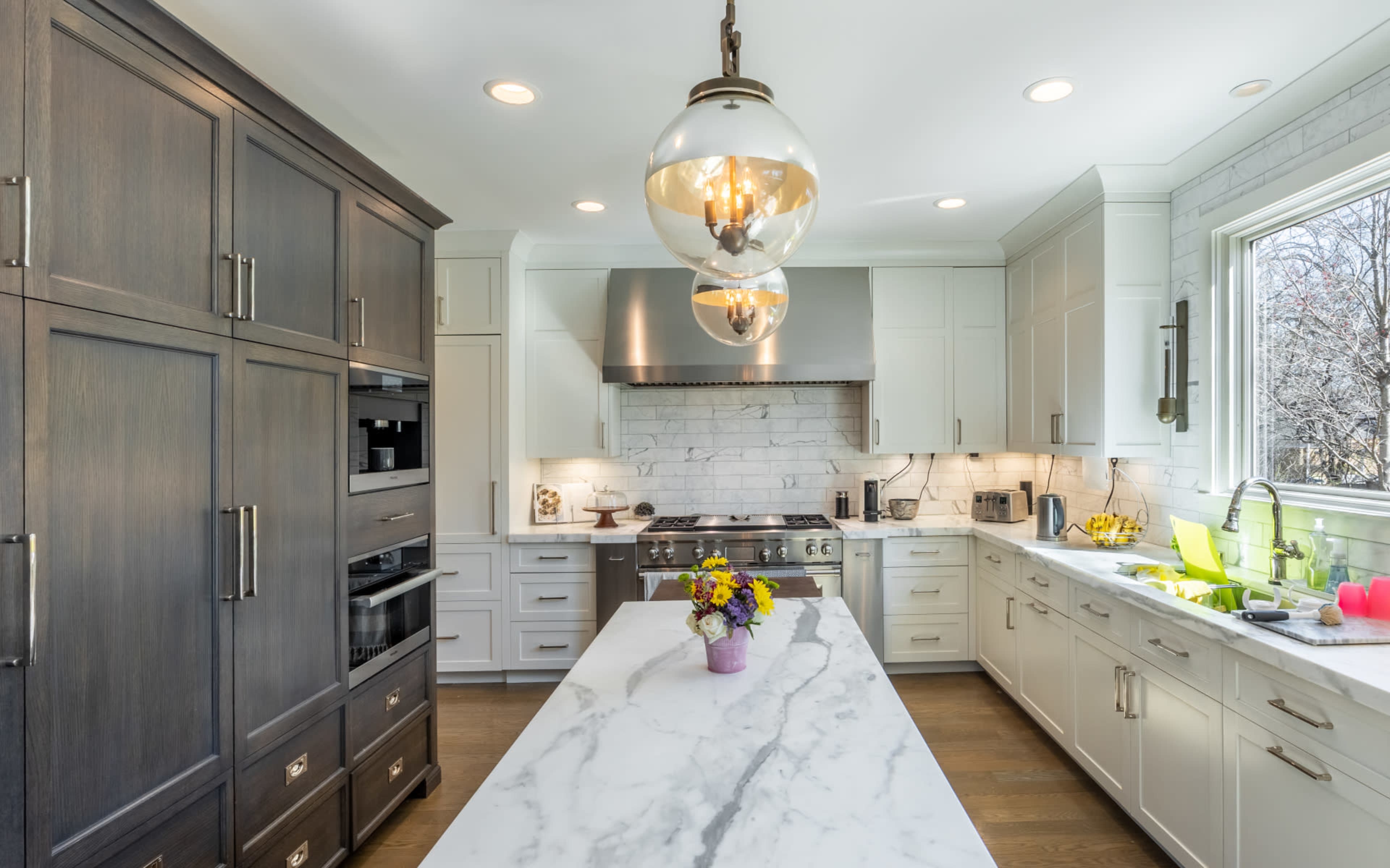 A modern kitchen features a large marble island, dark wood cabinets, stainless steel appliances, and a window overlooking a garden.