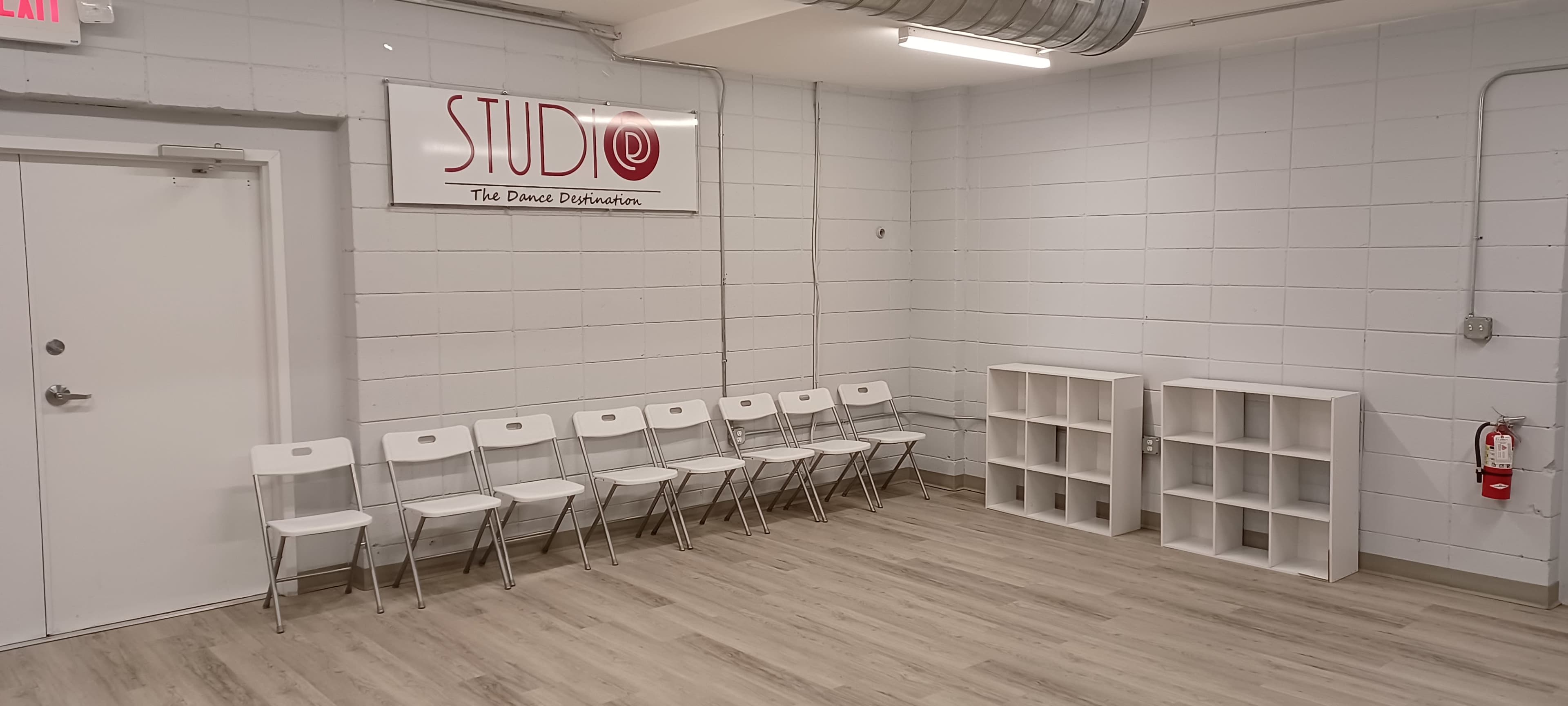 A room with a sign labeled "STUDIO" on the wall, featuring a row of white chairs and two shelving units.