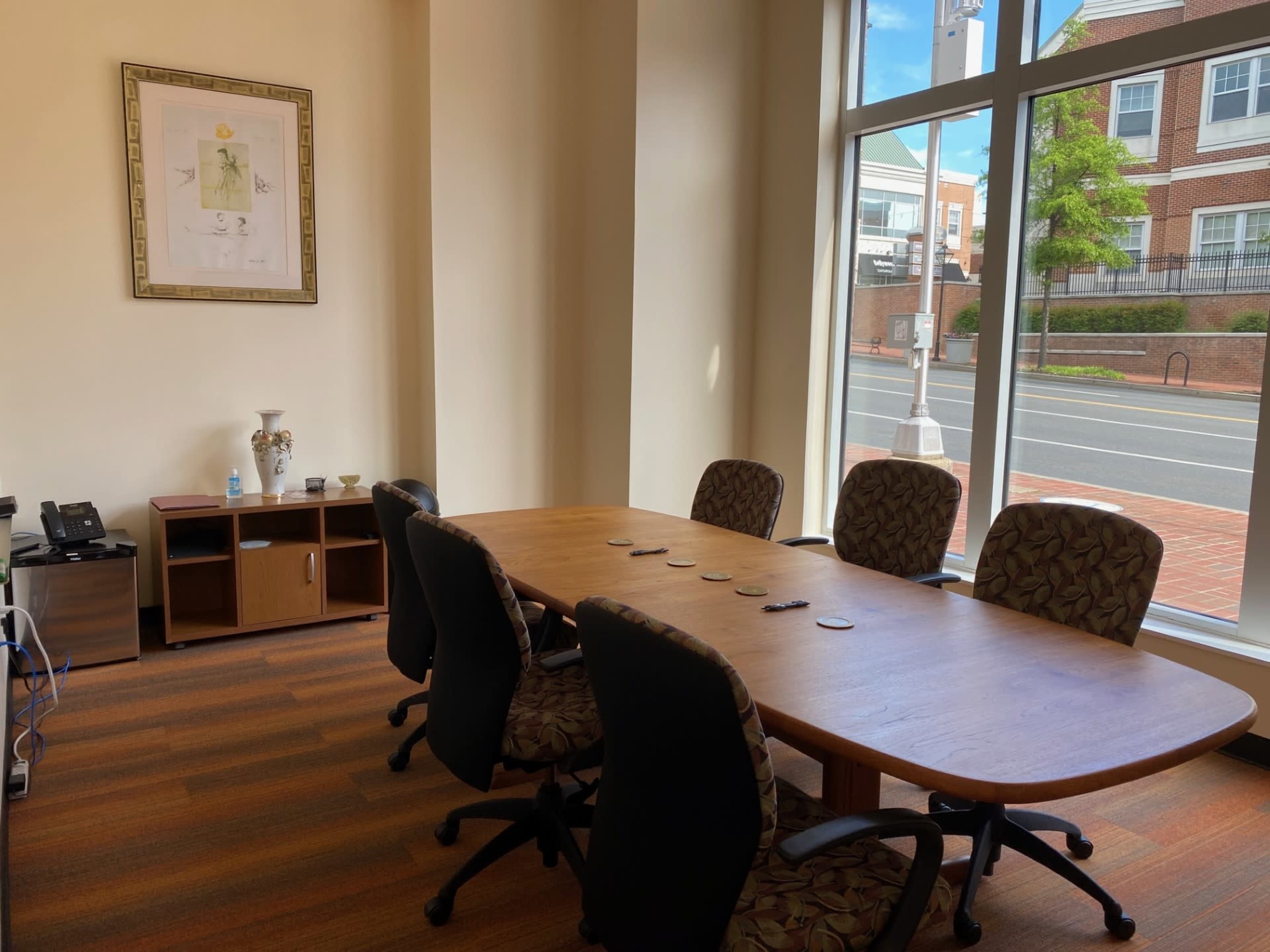 A conference room features a large wooden table surrounded by six patterned chairs, with a framed artwork on the wall and a view of the street outside.