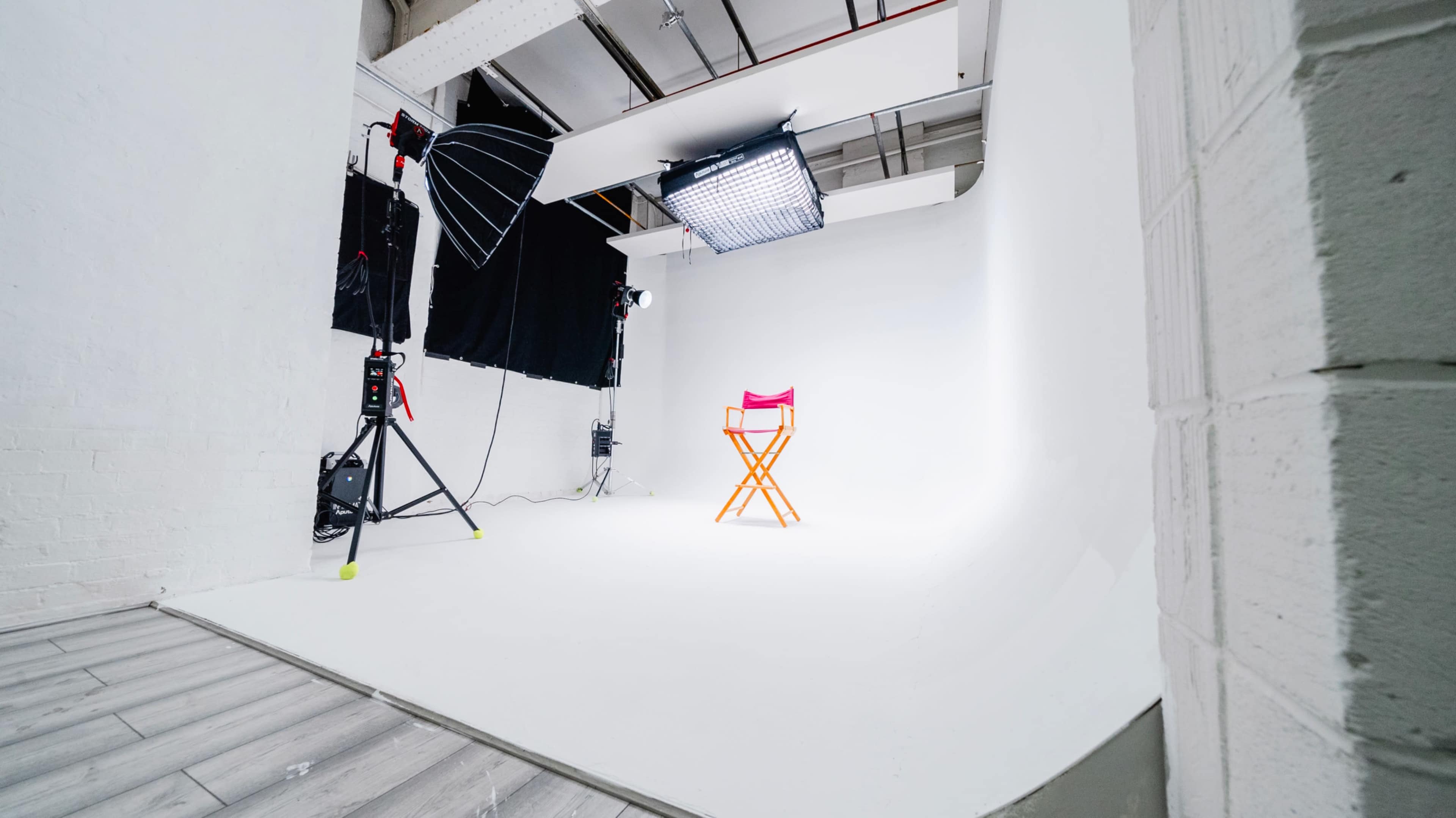The image shows a photography studio with a pink director's chair centered against a white backdrop, surrounded by lighting equipment.
