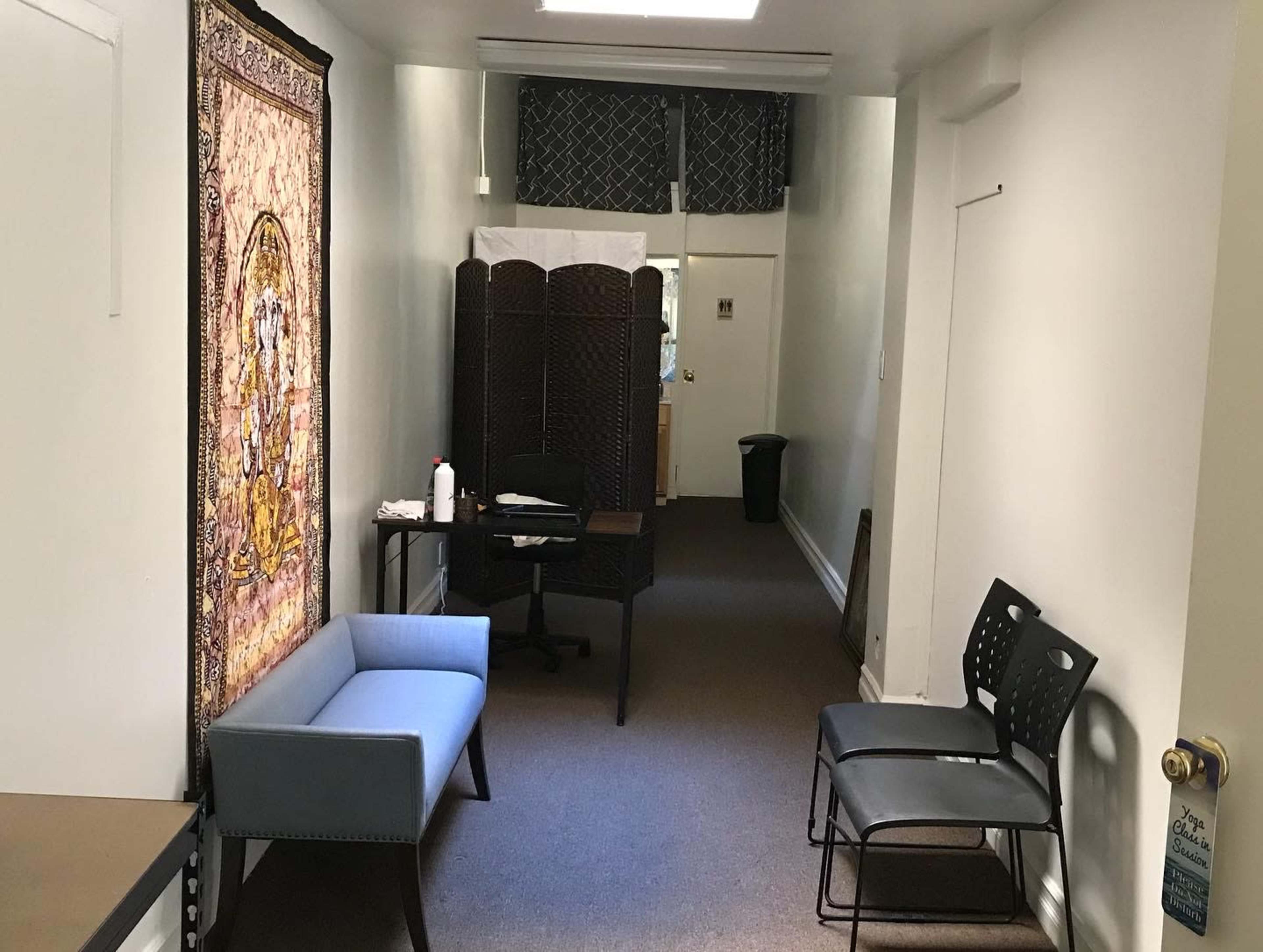 A narrow hallway with a tapestry on one wall, a seating area with two chairs and a small table, and a partially visible room at the end.