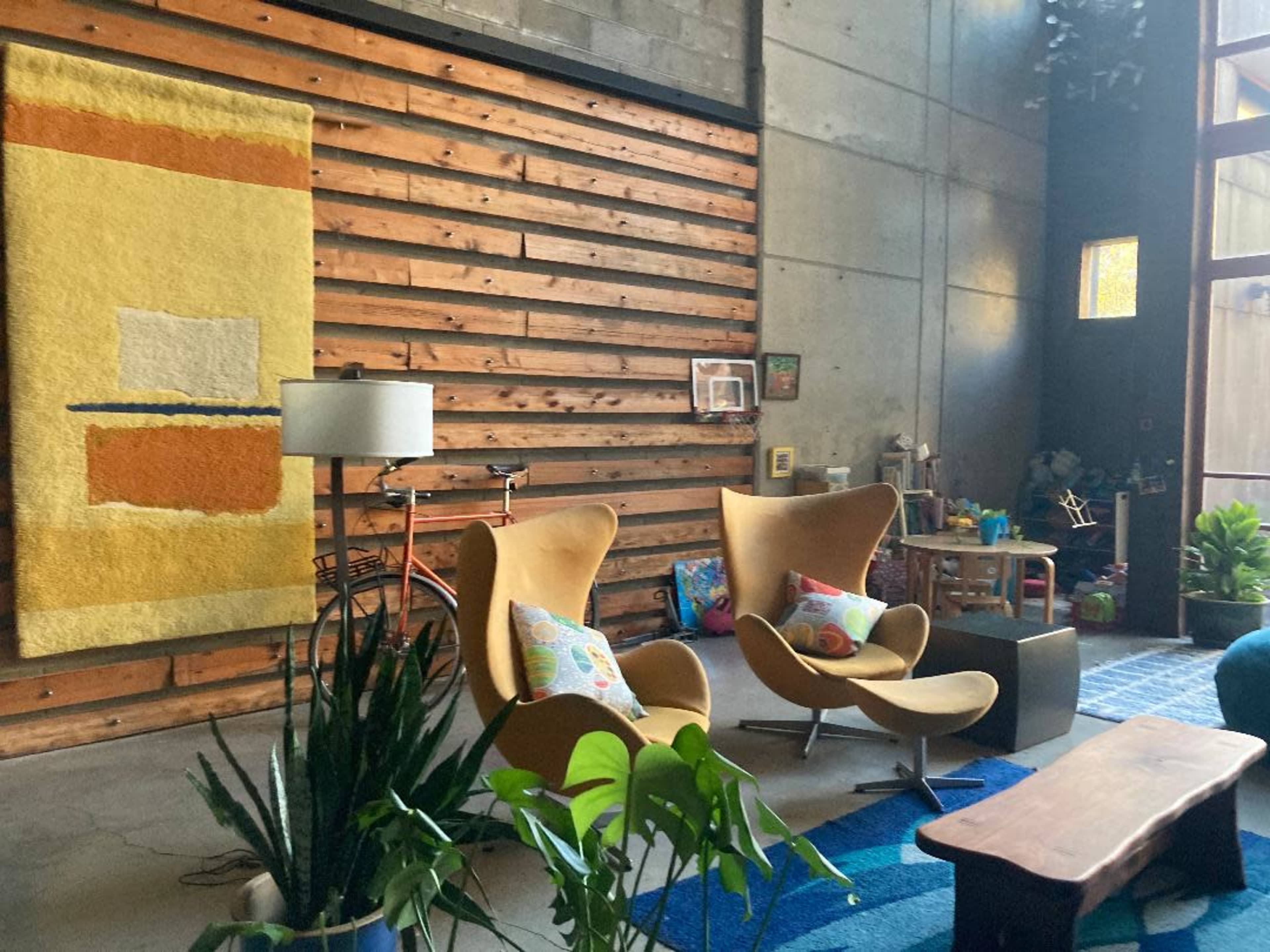 A modern living room featuring two yellow chairs, a wooden wall, a colorful textile art piece, and various plants.