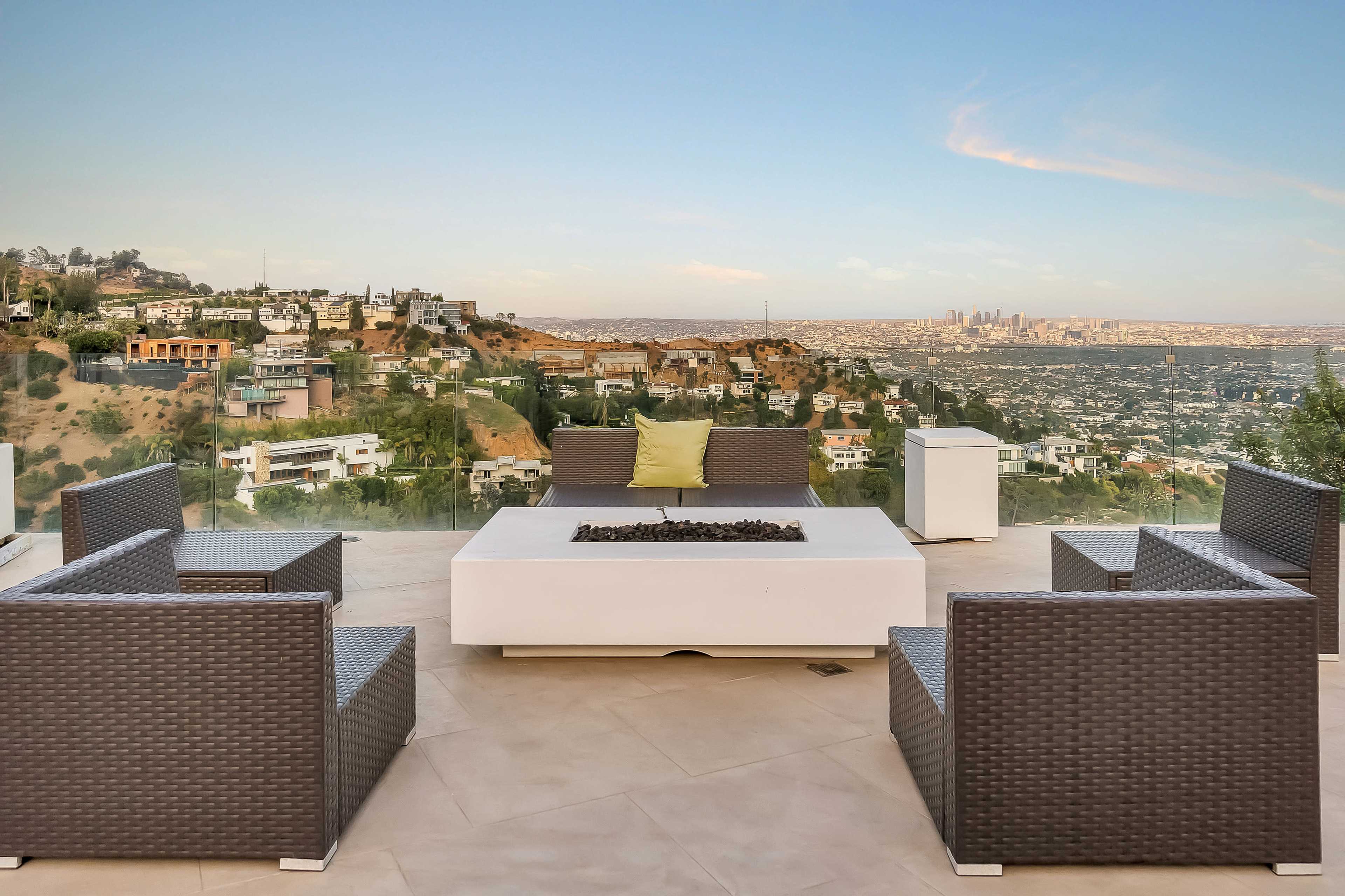 A modern outdoor seating area features a fire pit surrounded by wicker chairs, overlooking a panoramic view of a sprawling cityscape in the distance.