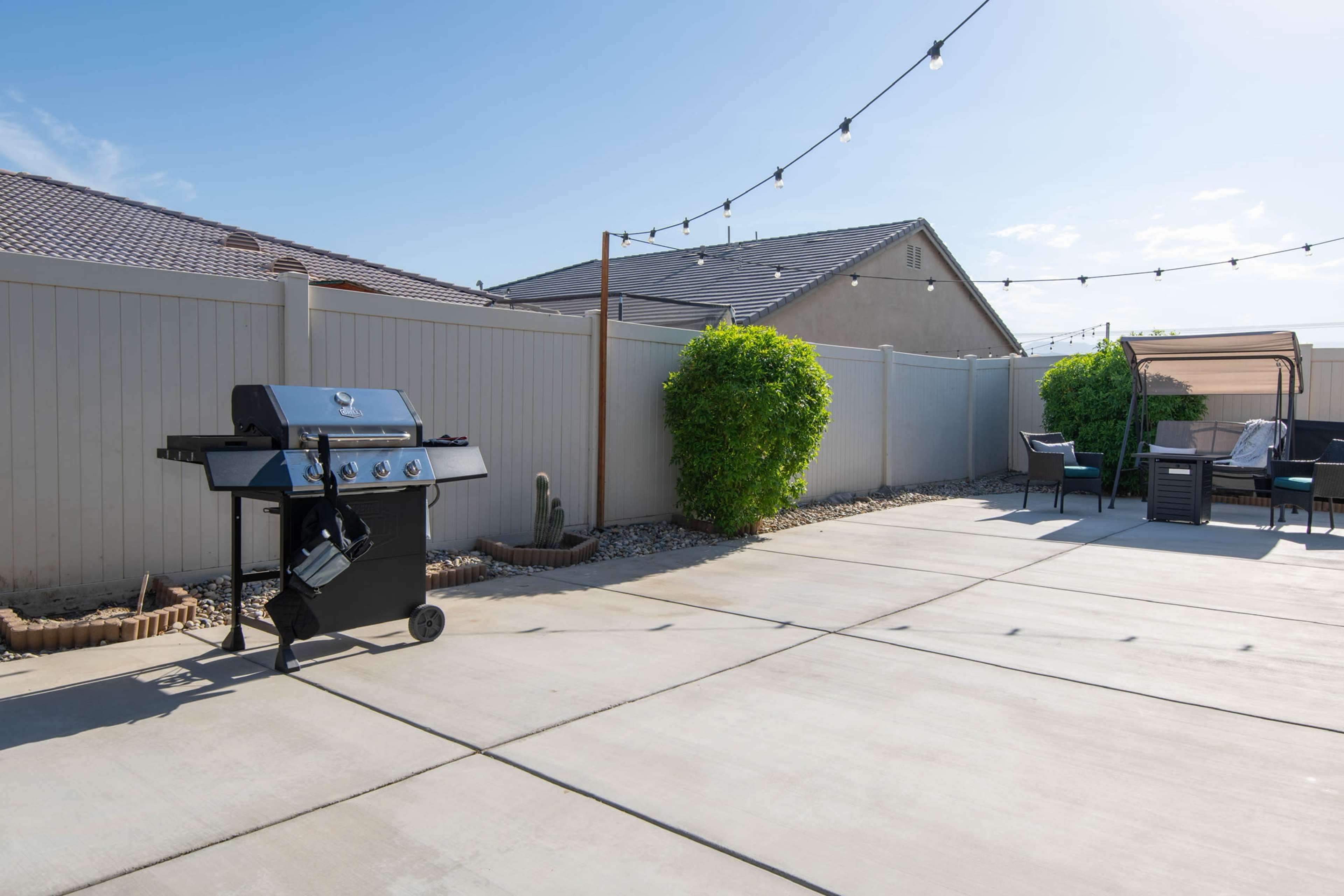 A backyard features a barbecue grill on a concrete patio surrounded by low shrubs and outdoor seating under string lights.