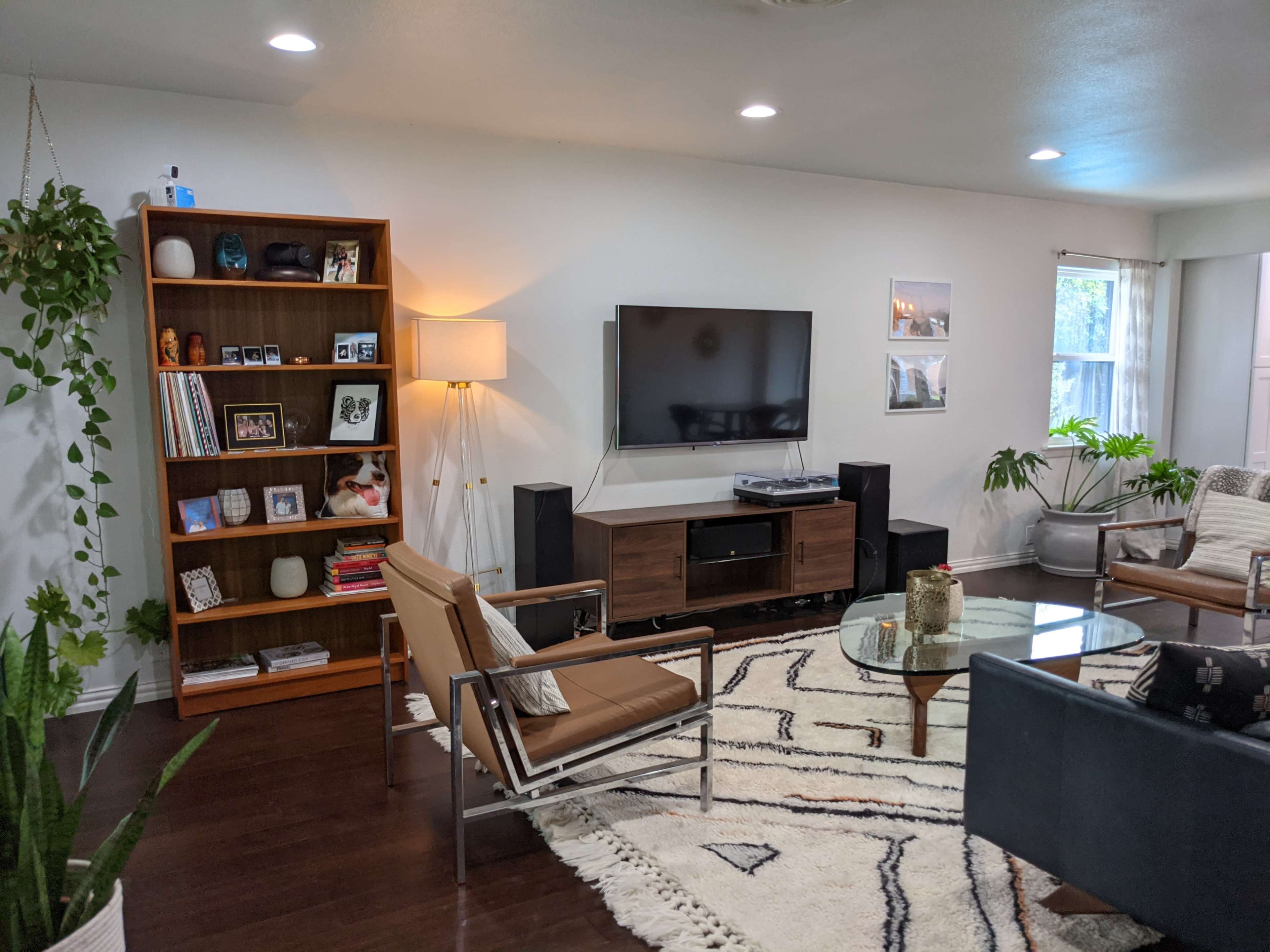 The living room features a bookshelf filled with books and decorative items, a television mounted on the wall, a coffee table in the center, and various plants scattered throughout the space.