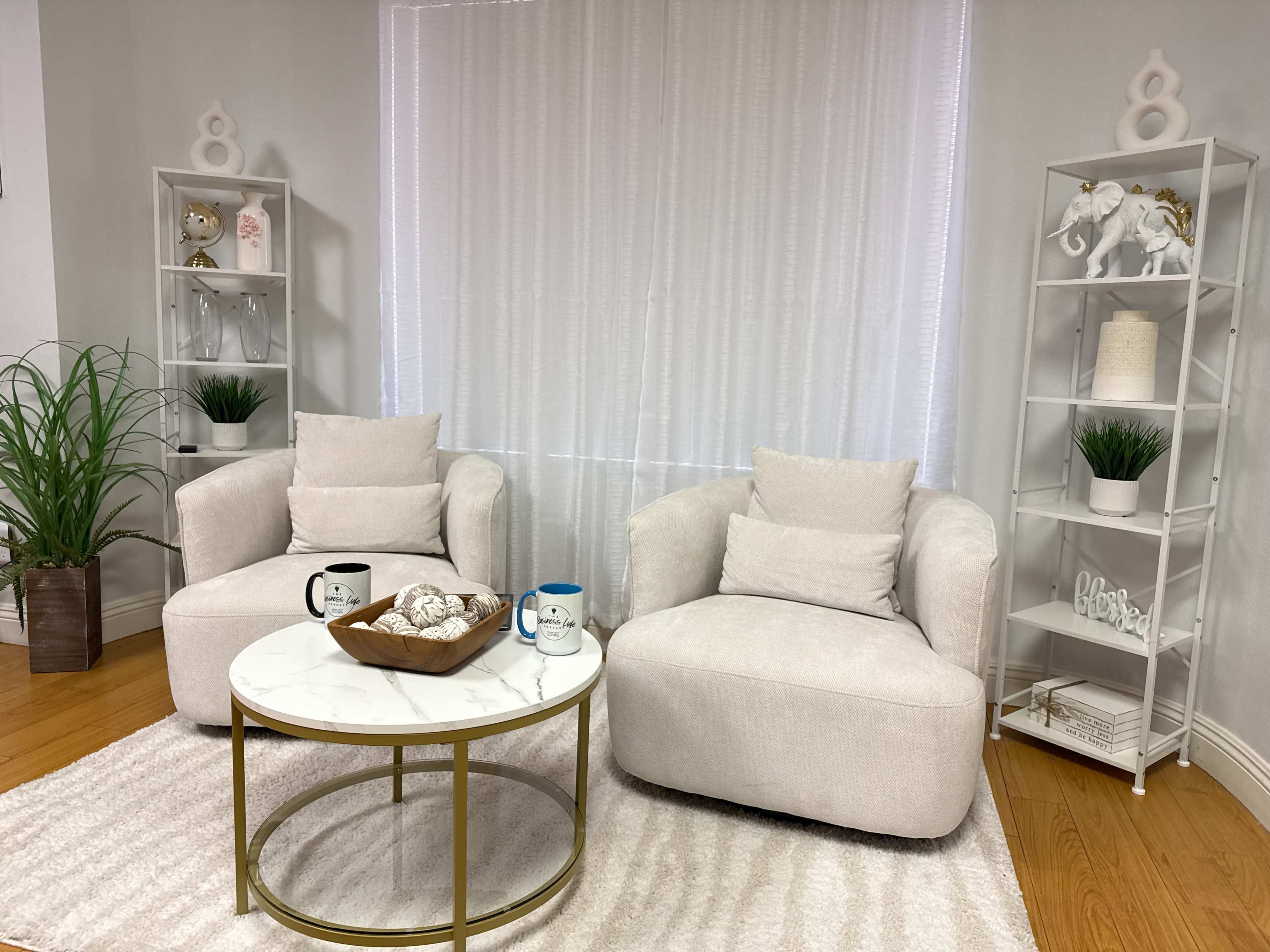 The image shows a modern living room setup with two plush armchairs, a round marble-topped coffee table, and decorative shelves featuring plants and ornaments.