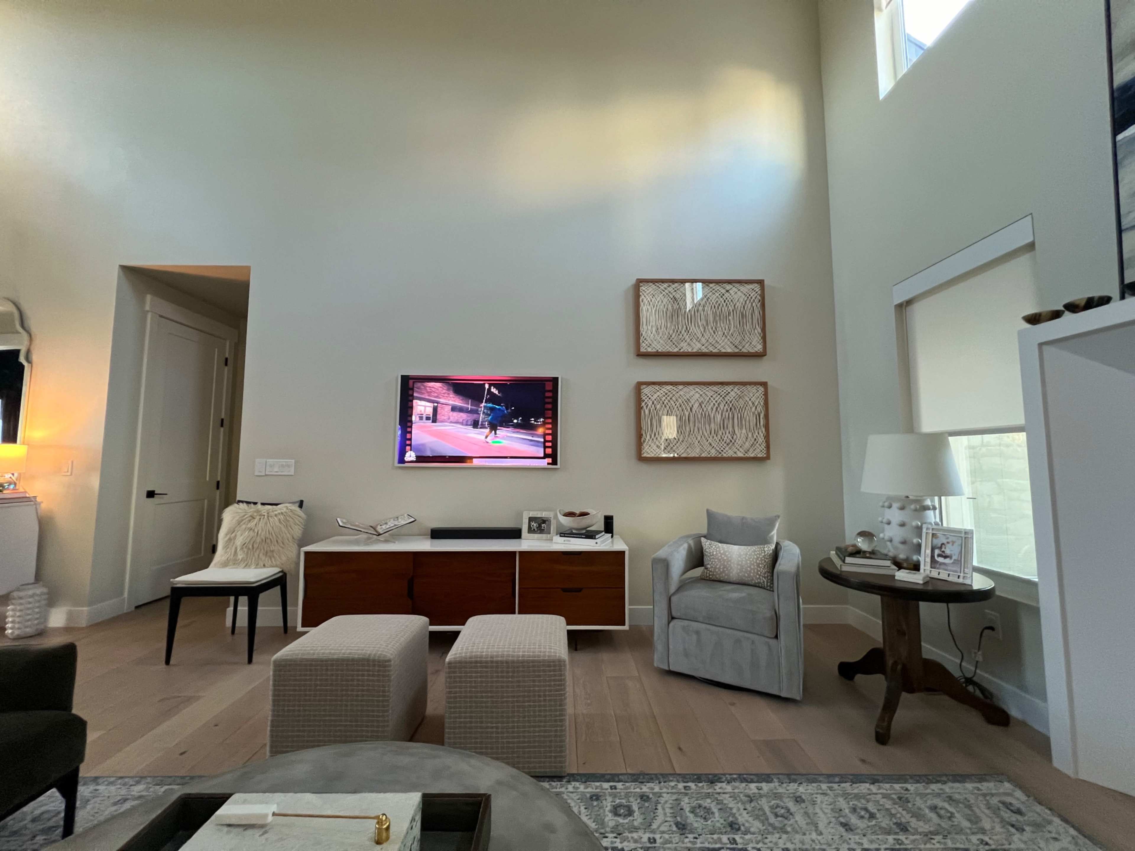 The image shows a modern living room with a wall-mounted television, a wooden console below it, two ottomans, and a chair in the corner beside a lamp.