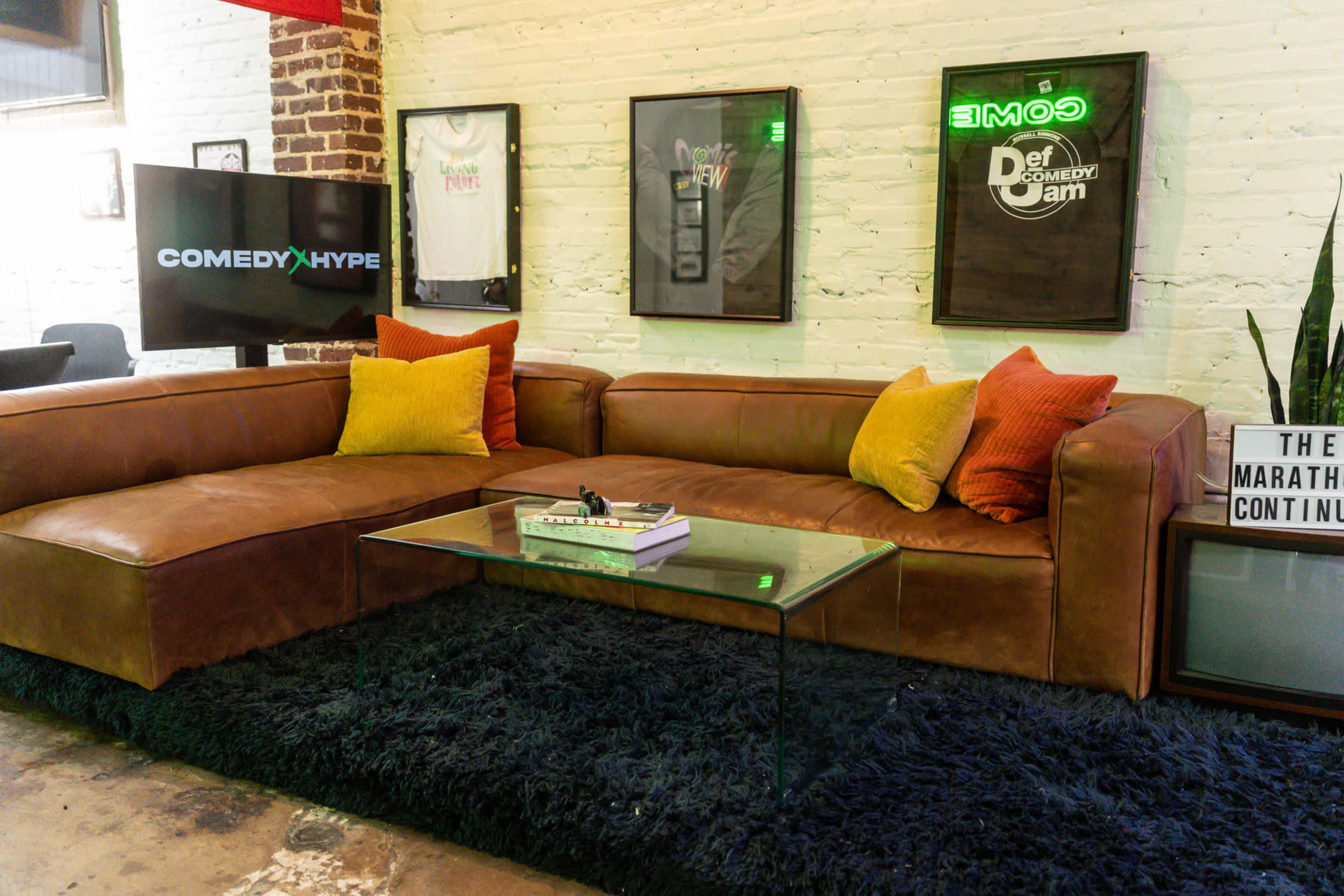 The image shows a modern lounge area featuring a large brown leather sectional sofa with colorful throw pillows, a glass coffee table, and framed posters on the wall.