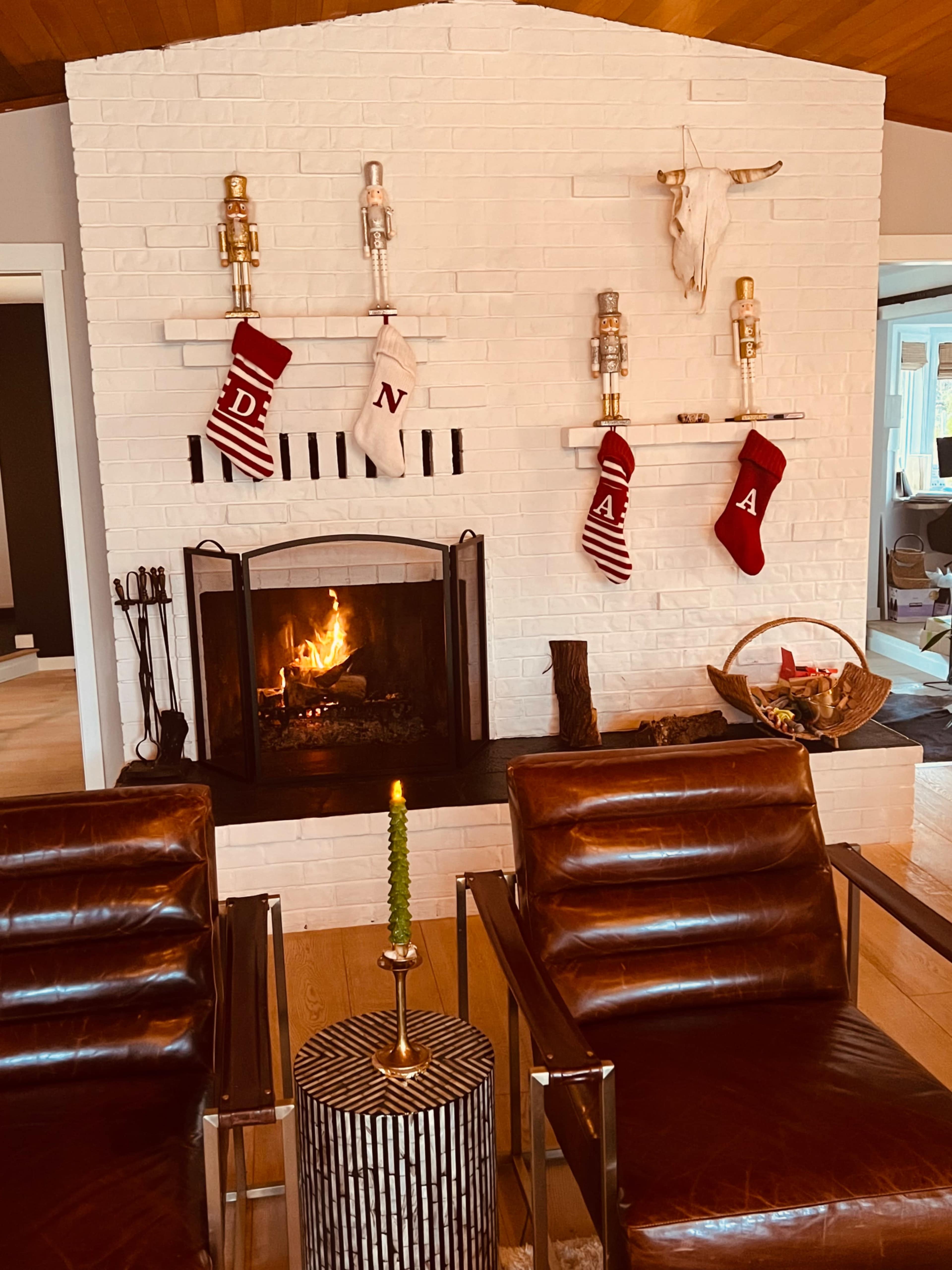A cozy living room features a white brick fireplace adorned with holiday stockings and a lit fire, flanked by two brown leather chairs and a decorative table.