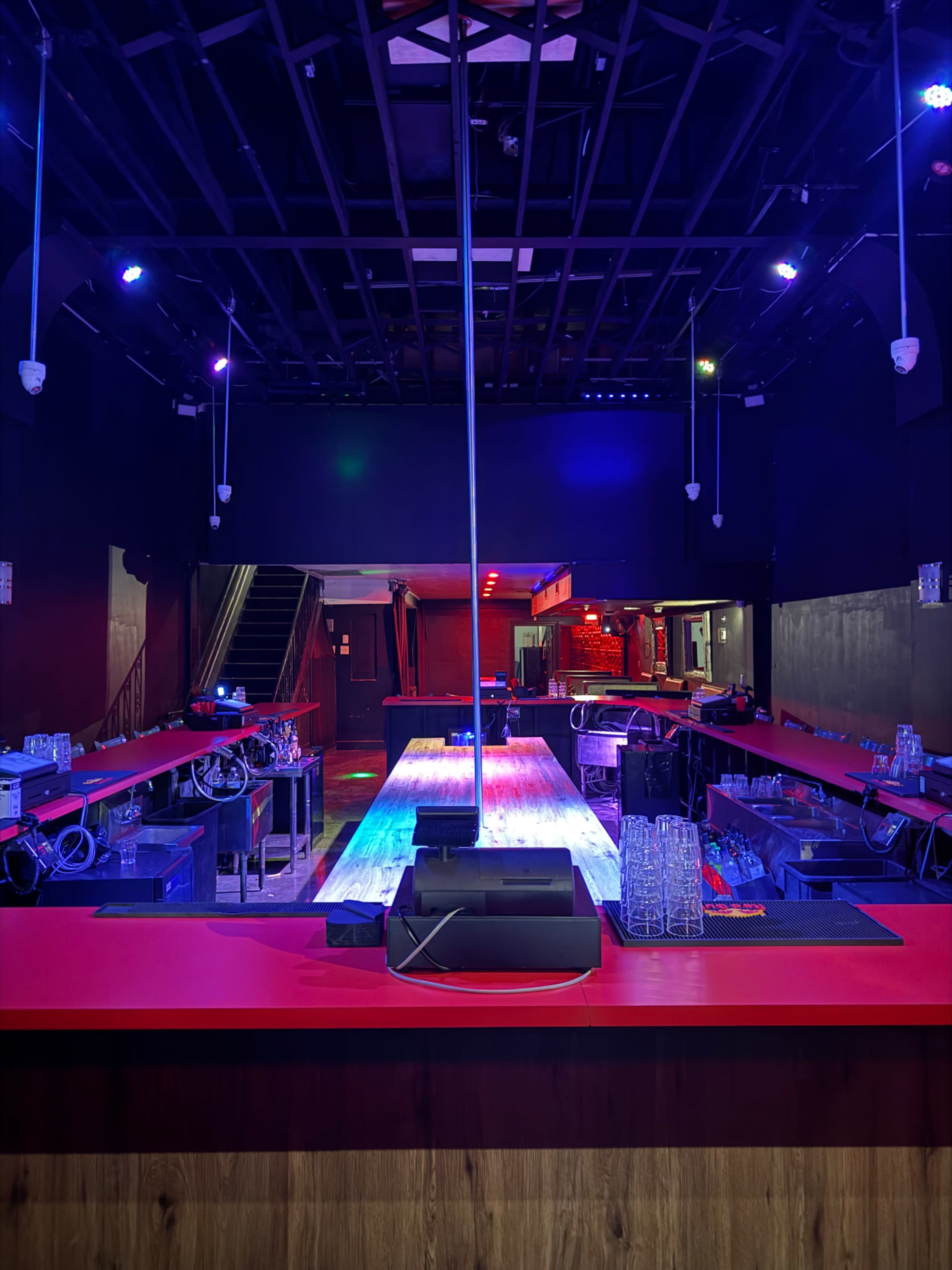 The image shows a dimly lit nightclub interior featuring a long red bar with a central pole, illuminated by colorful lights and flanked by shelves and glassware.
