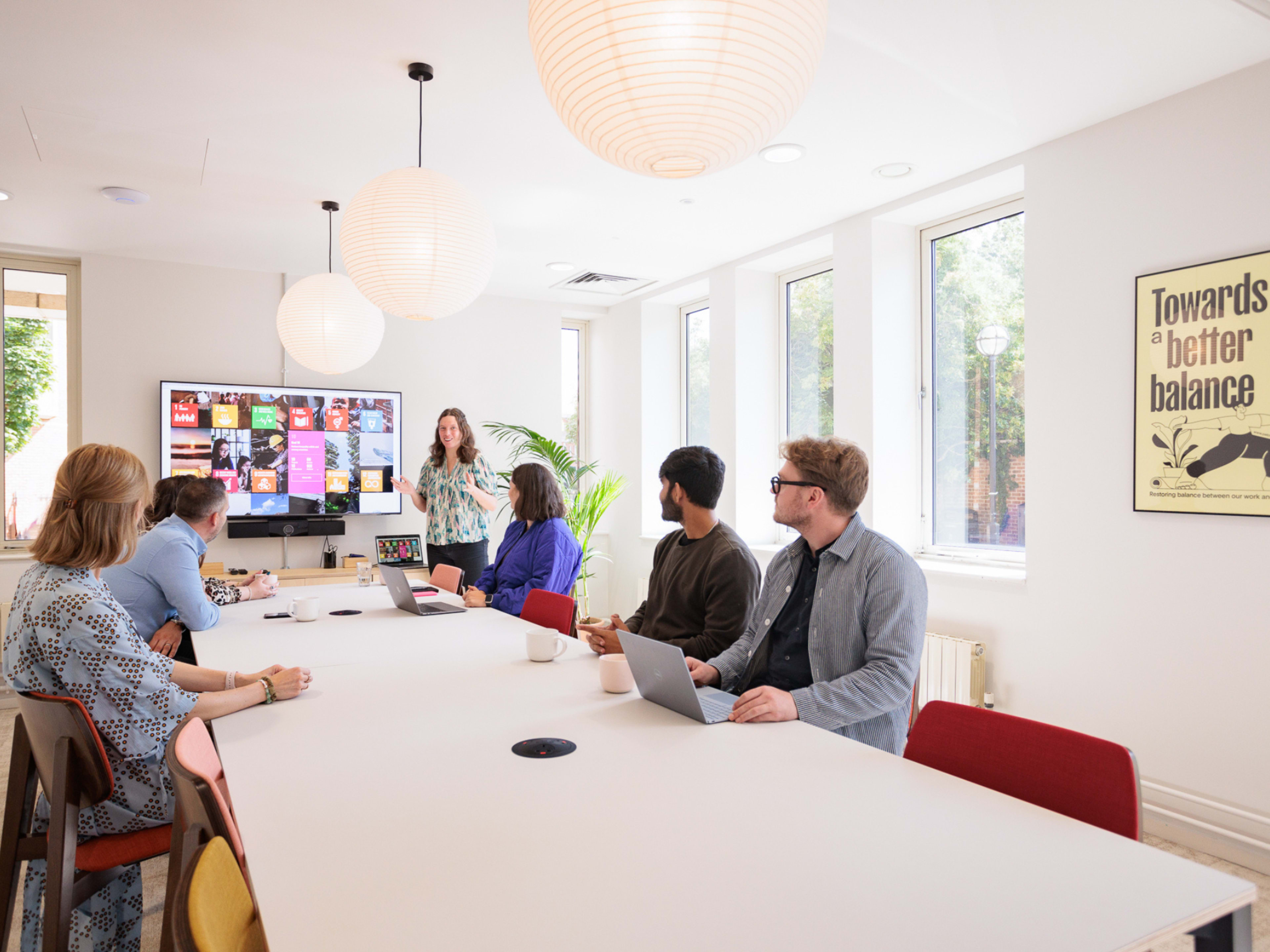 A group of six people is seated around a long table in a bright meeting room, with one person presenting on a screen at the front.