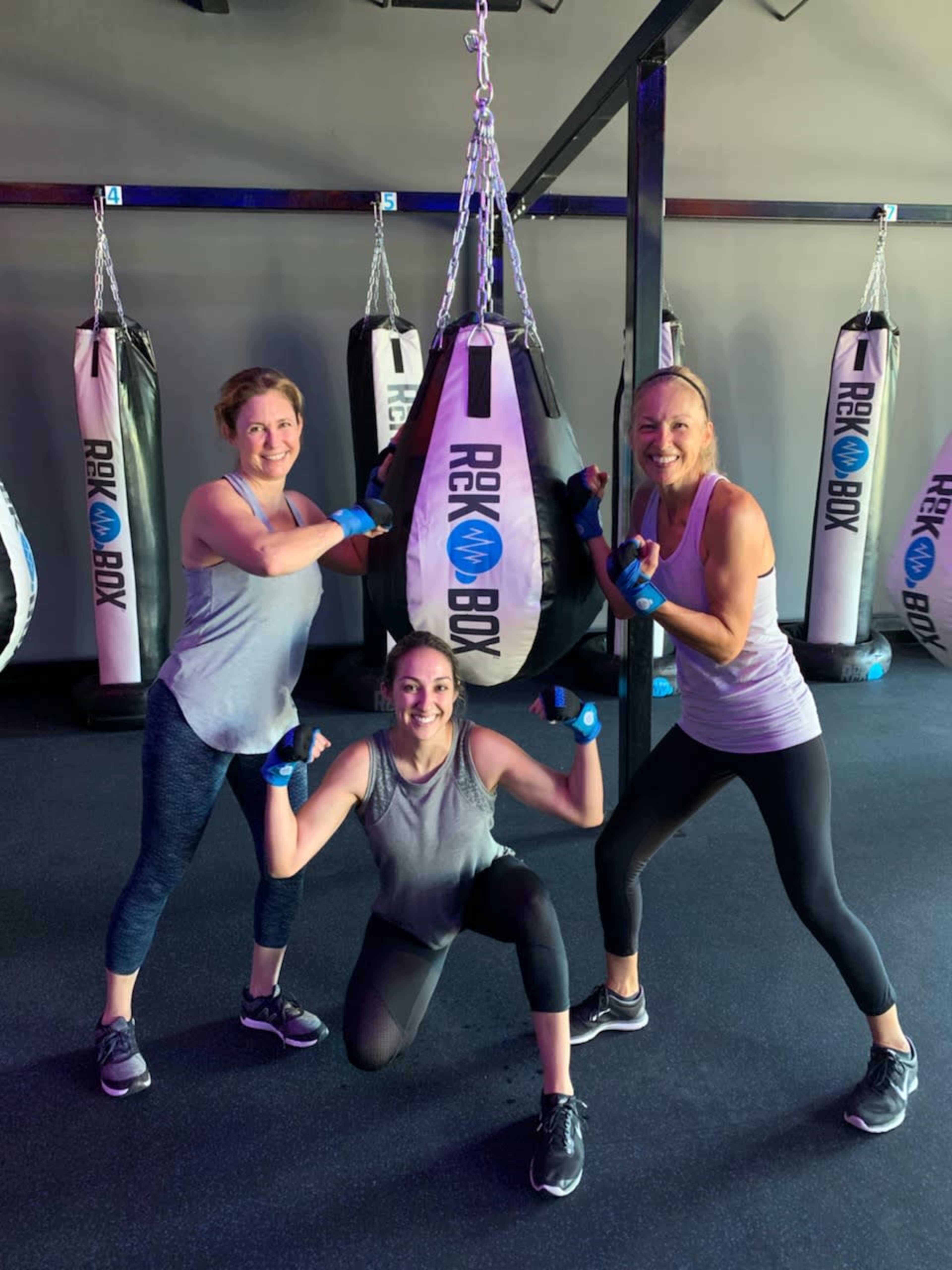 Three women pose playfully in a gym with punching bags in the background.