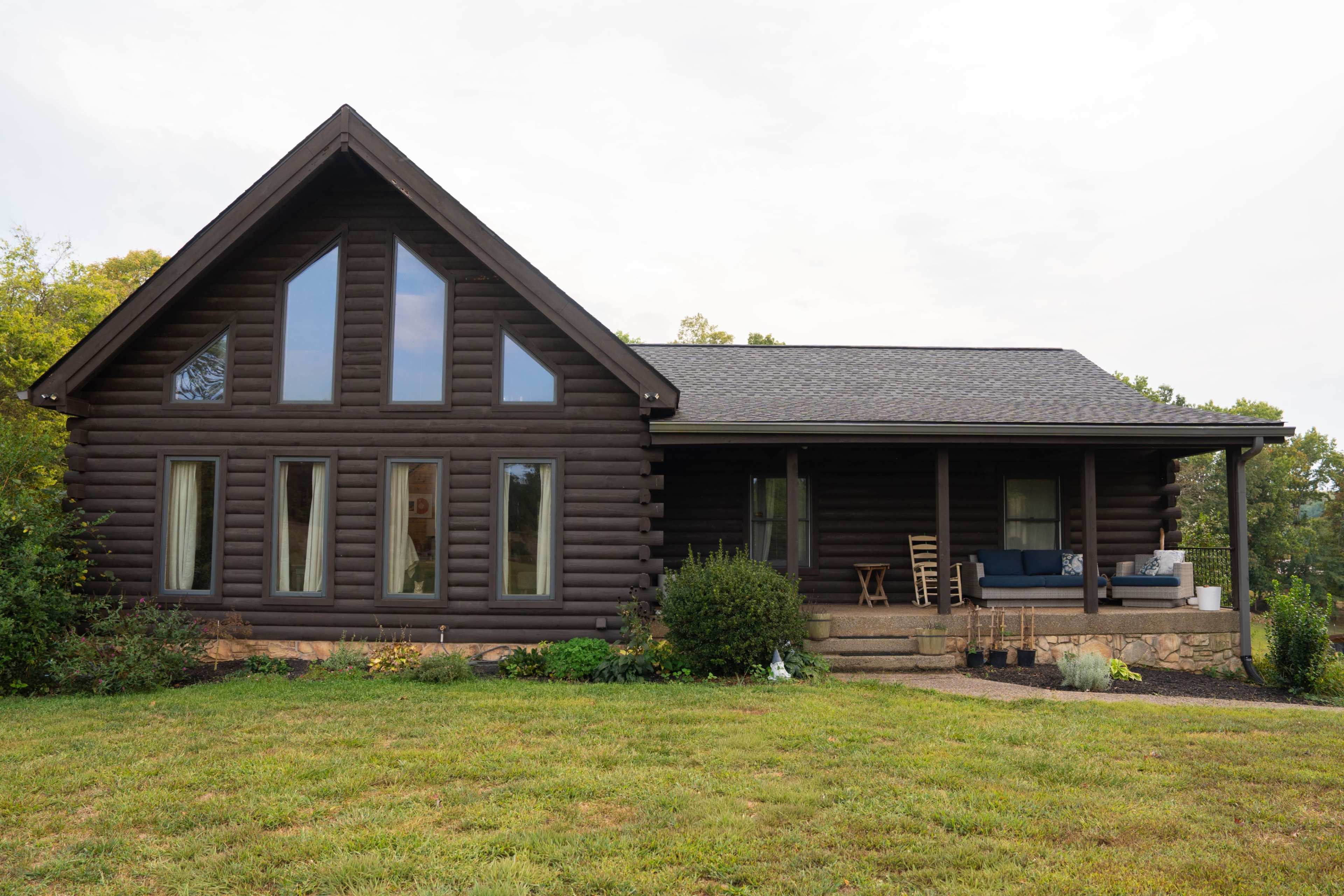 The image features a rustic log cabin with a sloped roof, large front windows, and a porch with seating.