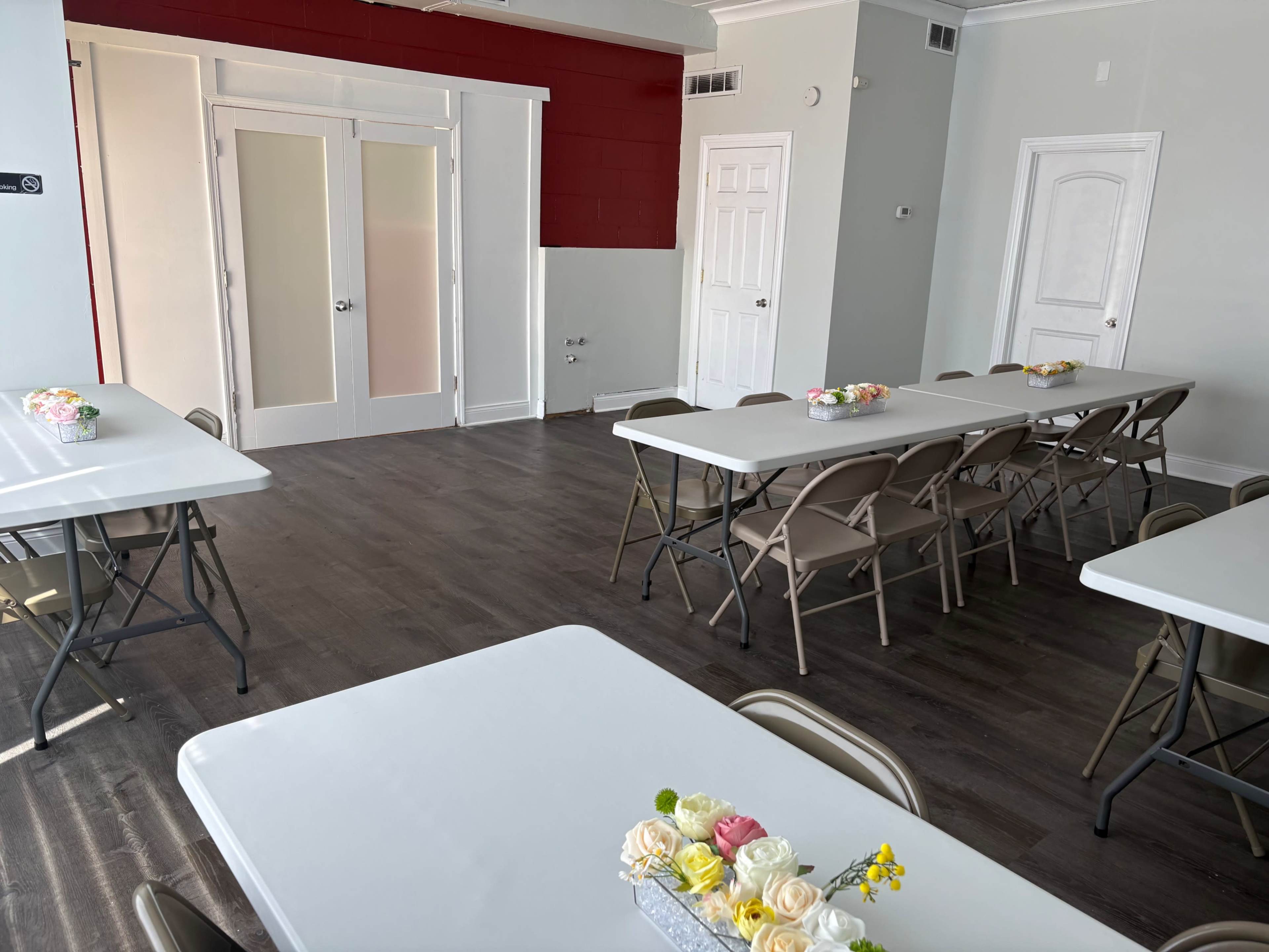 The image shows a bright room with several folding tables arranged in rows, each decorated with small flower centerpieces, and a red accent wall behind a pair of double doors.