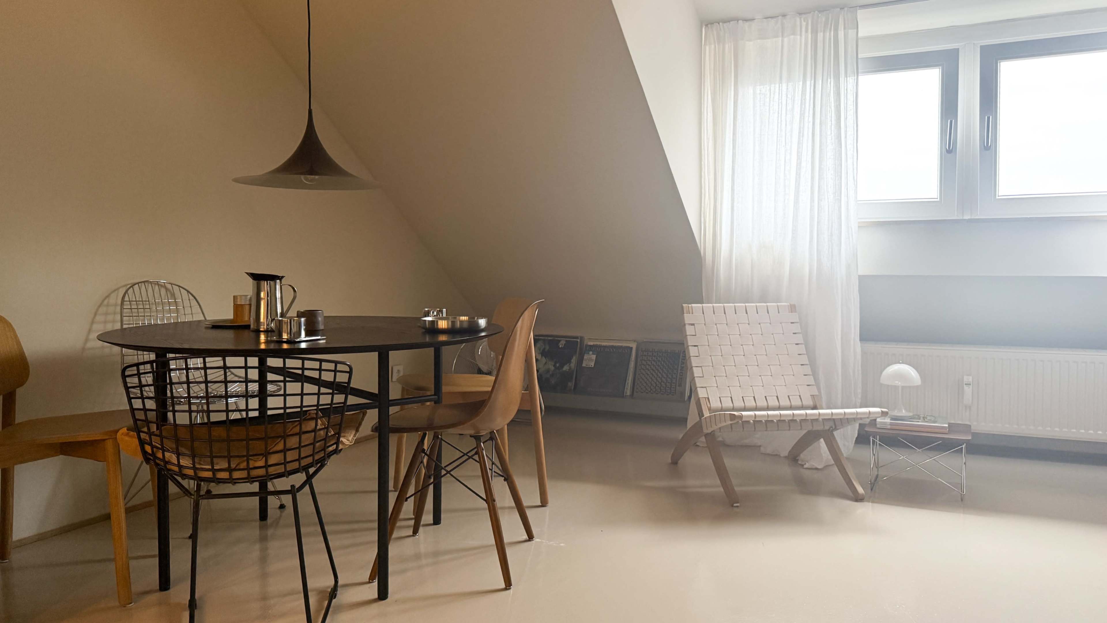 A cozy dining area features a round table with wire chairs, a pendant light overhead, and a comfortable lounge chair beside large windows adorned with sheer curtains.